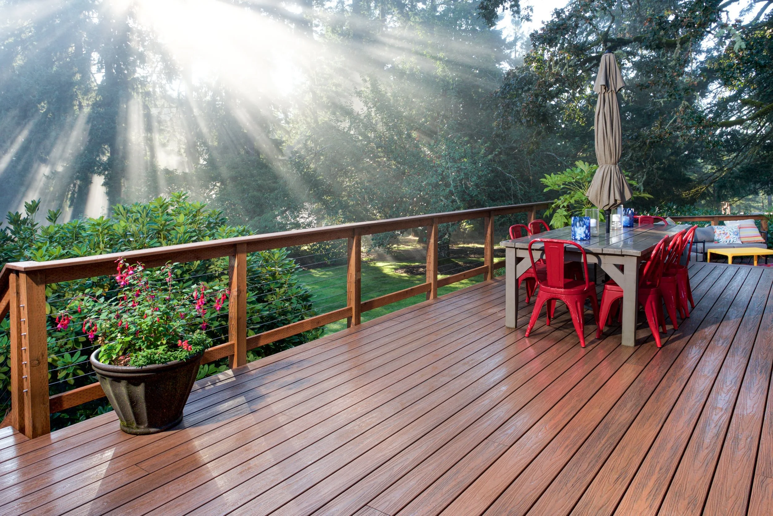 A wooden deck with a large dining table and red chairs, a closed patio umbrella, lush green trees, sunlight streaming through the trees, and potted plants with pink flowers.