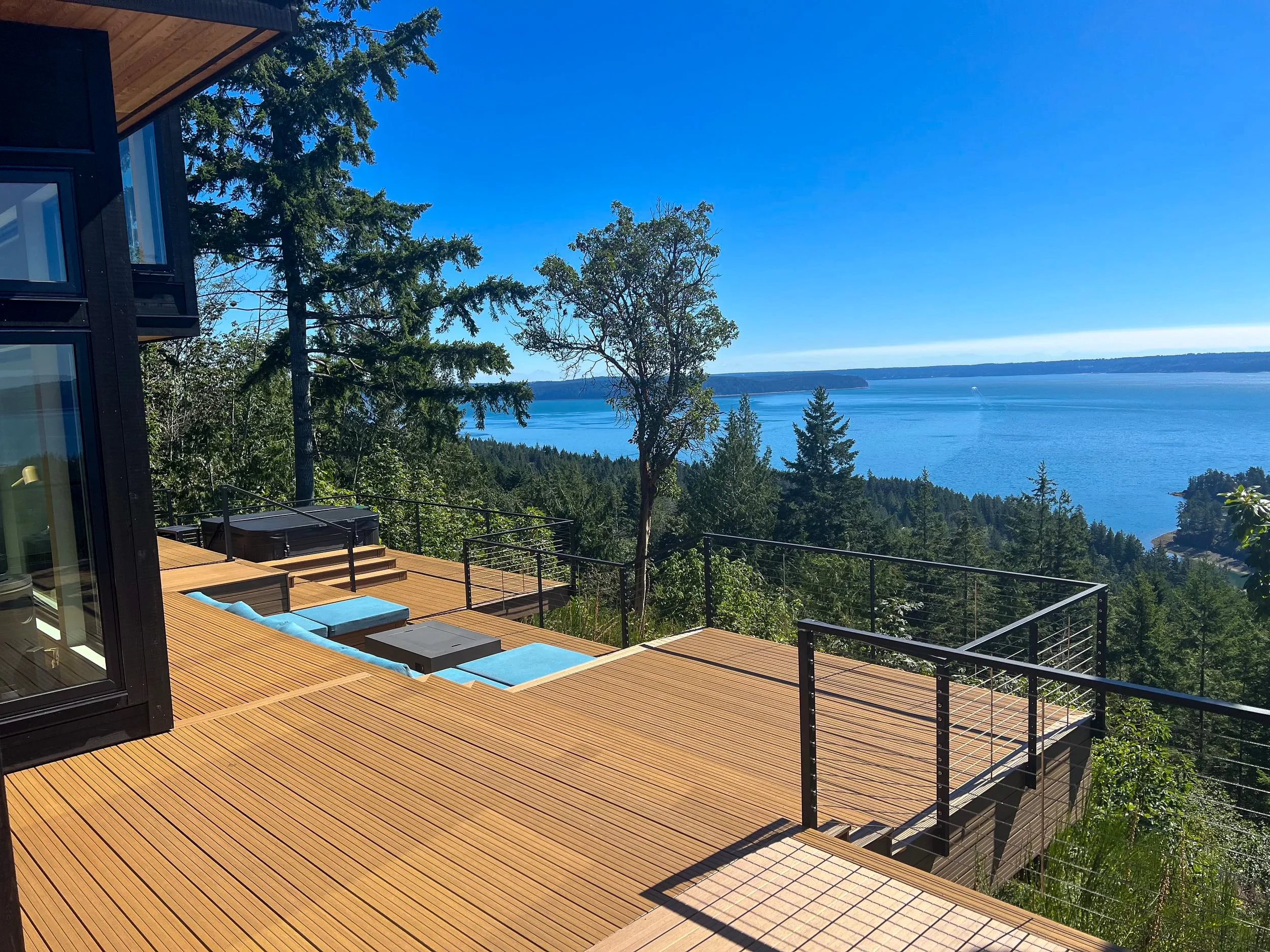A spacious wooden deck overlooking a lake, surrounded by trees, with a partial view of a modern building and outdoor furniture.