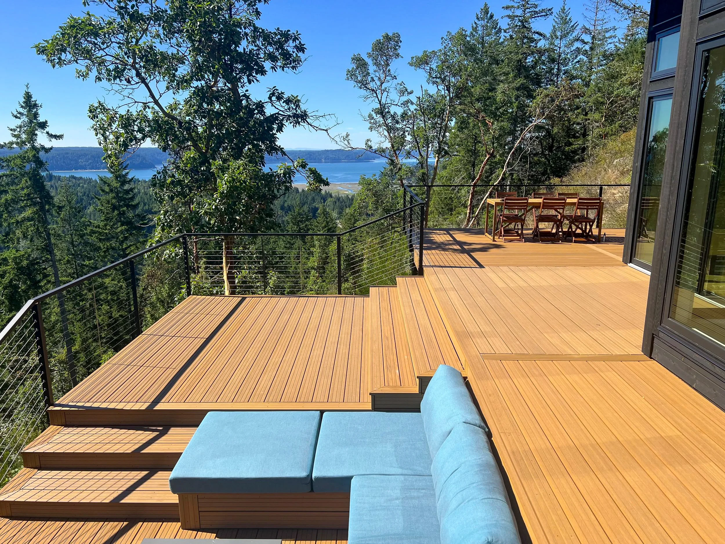 View of a spacious wooden deck overlooking a lush forest and a distant lake with blue sky above. There is a seating area with blue cushions and a wooden dining table with chairs.