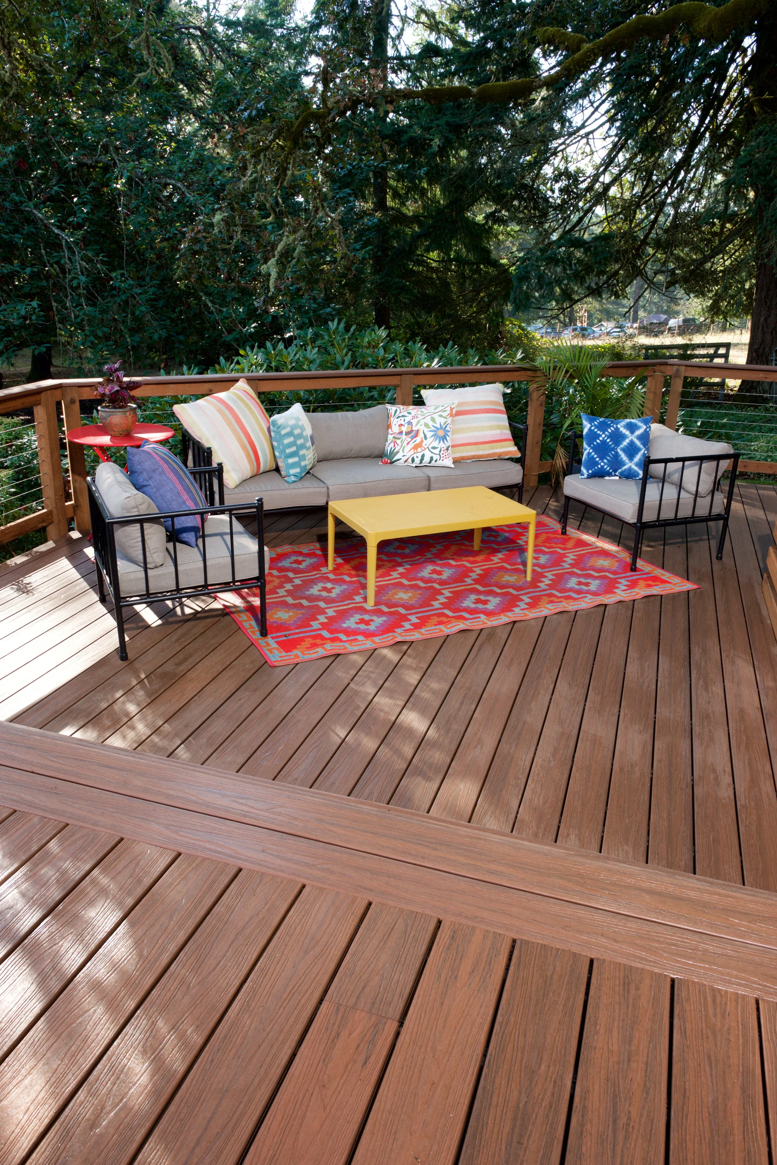 A wooden outdoor deck with a couch, chairs, colorful pillows, a yellow coffee table, a red side table, and a patterned rug, surrounded by greenery and trees.