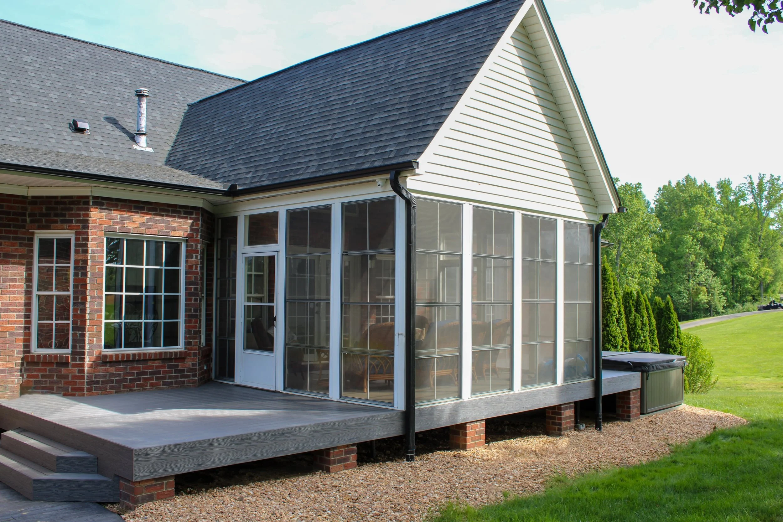 A house with a red brick exterior and a large screen porch with metal and glass panels, a grey wooden deck, and a shingled roof with a vent pipe and a chimney.