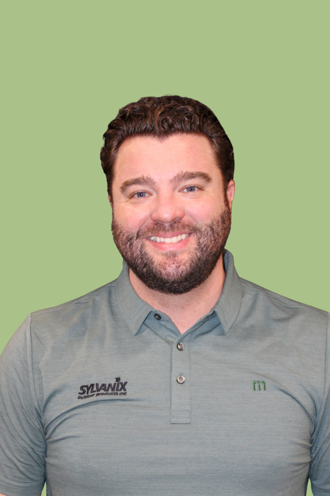 A smiling man with brown hair and a beard, wearing a gray collared shirt with logos, standing against a light green background.