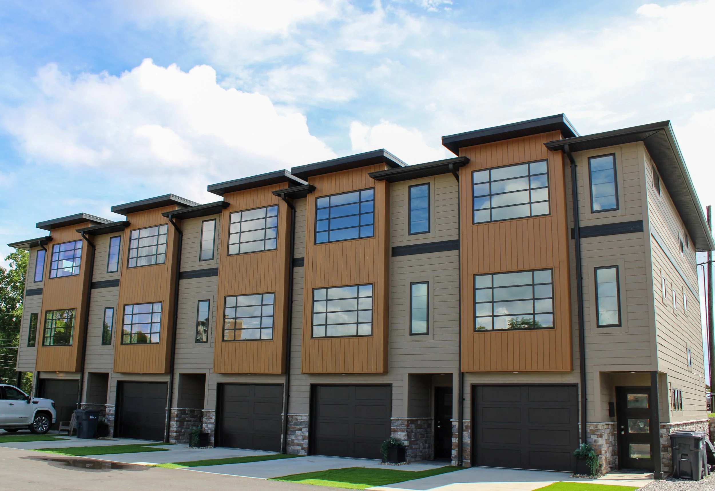Modern multi-story apartment building with large windows, garage doors at ground level, a blue sky with clouds, parked cars, and greenery in front.