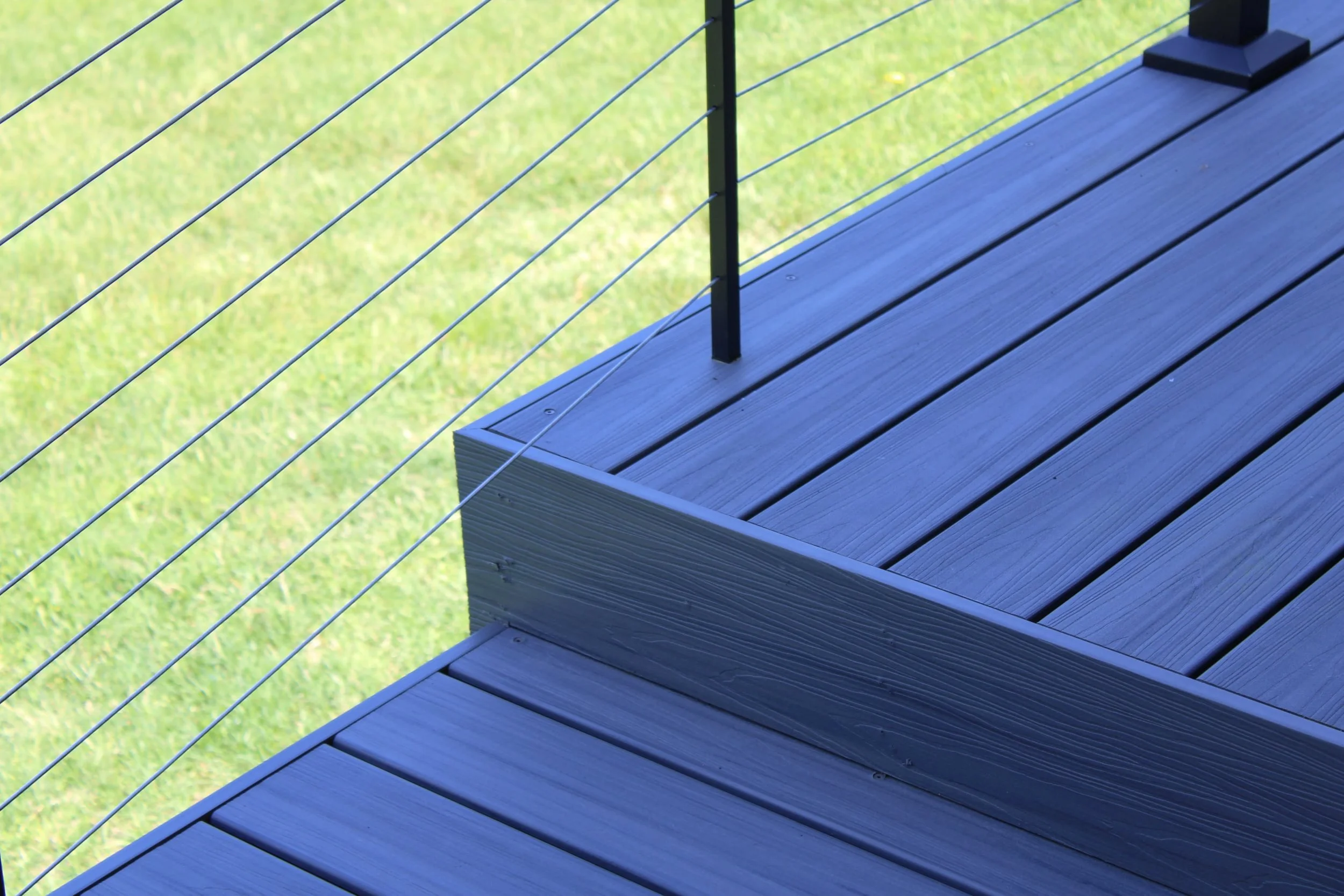 Close-up view of a wooden deck with metal cable railing and green grass background.