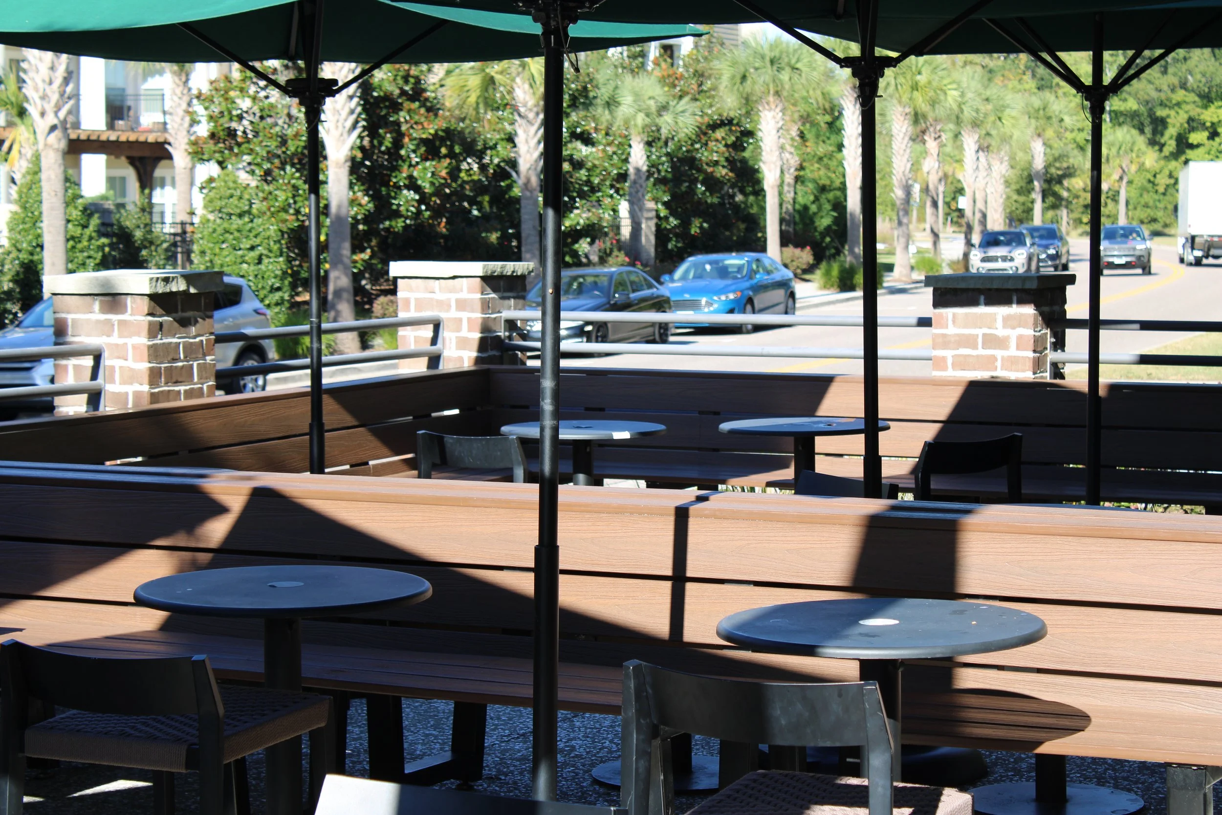 Outdoor seating area with round tables and chairs, shaded by large umbrellas, overlooking a street with parked cars, trees, and a building in the background.