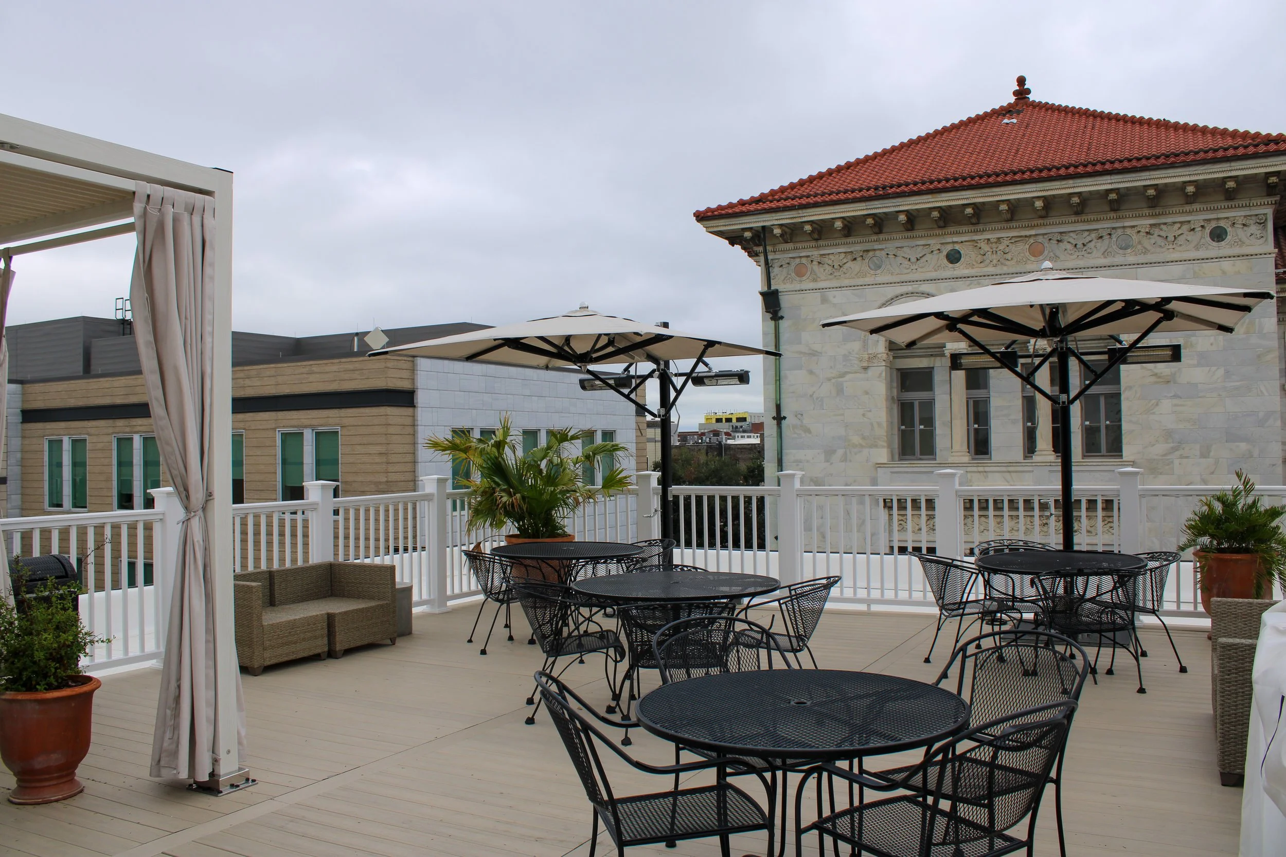 An outdoor rooftop patio with black metal tables and chairs, white umbrellas, potted plants, and a white railing. There is a historic building with a red tiled roof and intricate architectural details in the background, under a cloudy sky.