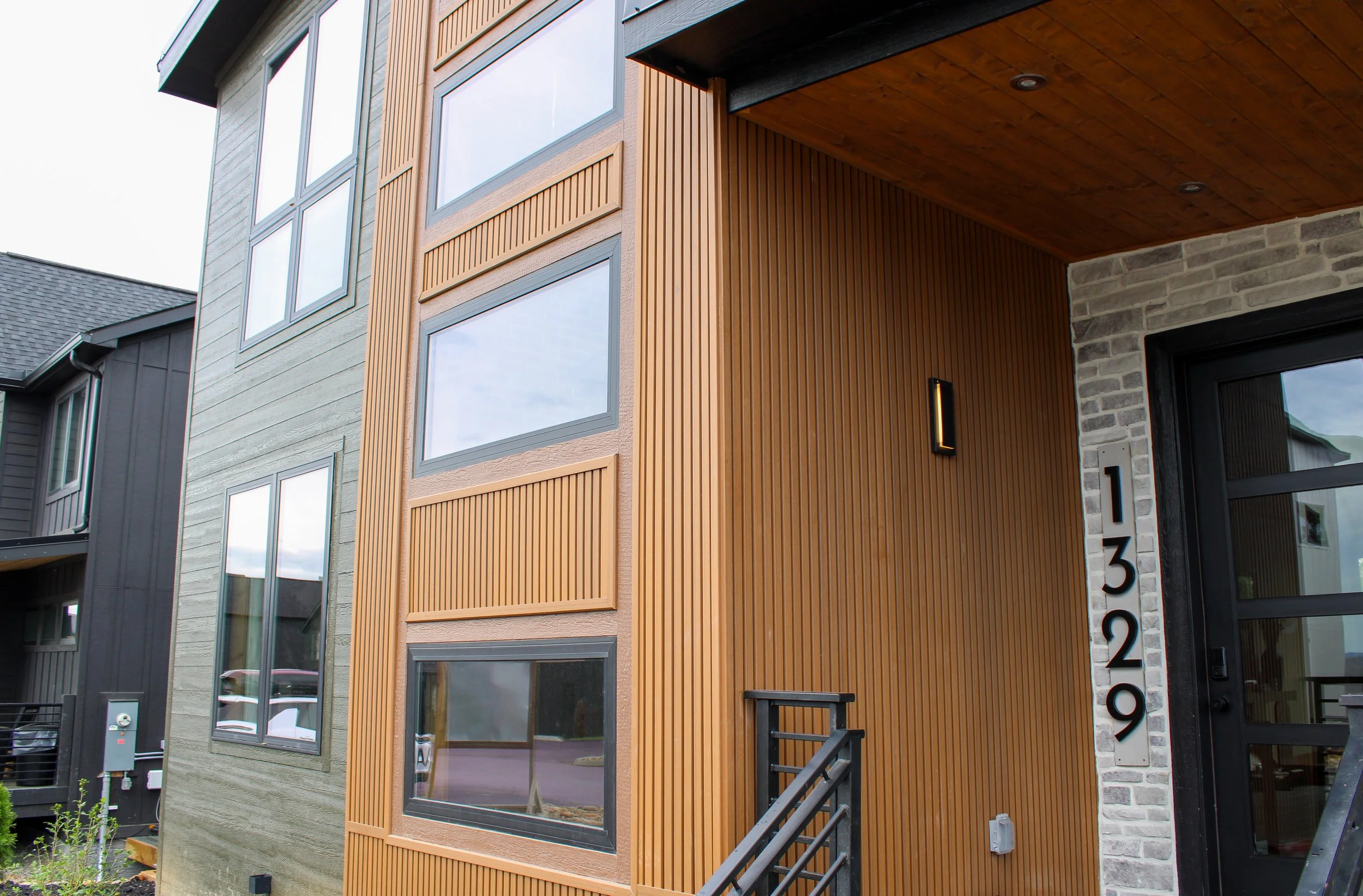 Close-up of a modern multi-story building with a facade combining gray horizontal siding, vertical wooden slats, and gray brickwork near the entrance. Large windows reflect the sky and neighboring structures, and there's a black exterior light beside the door with a house number display.