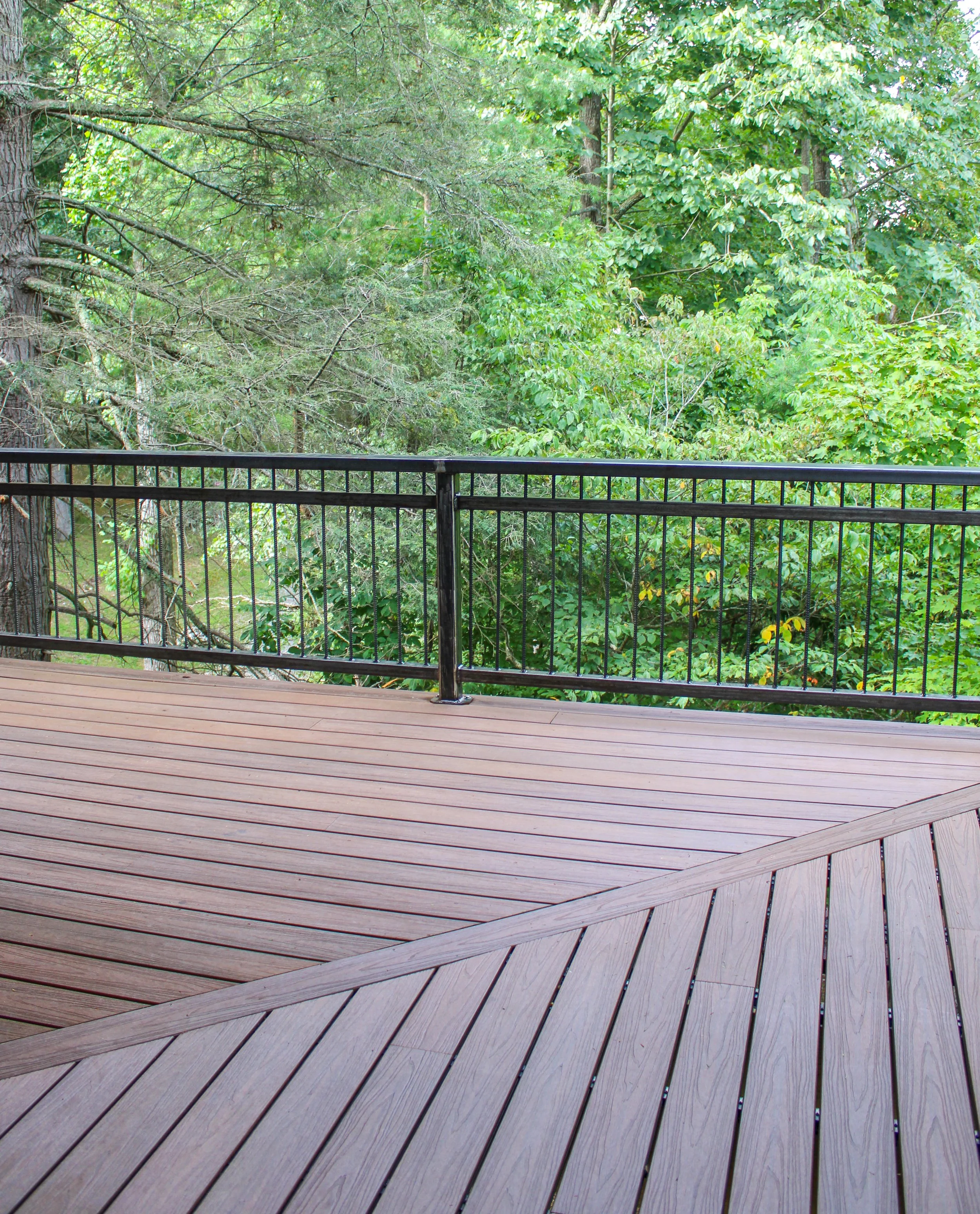 A wooden deck with curved and straight lines, bordered by a black metal railing, overlooking lush green trees and foliage.