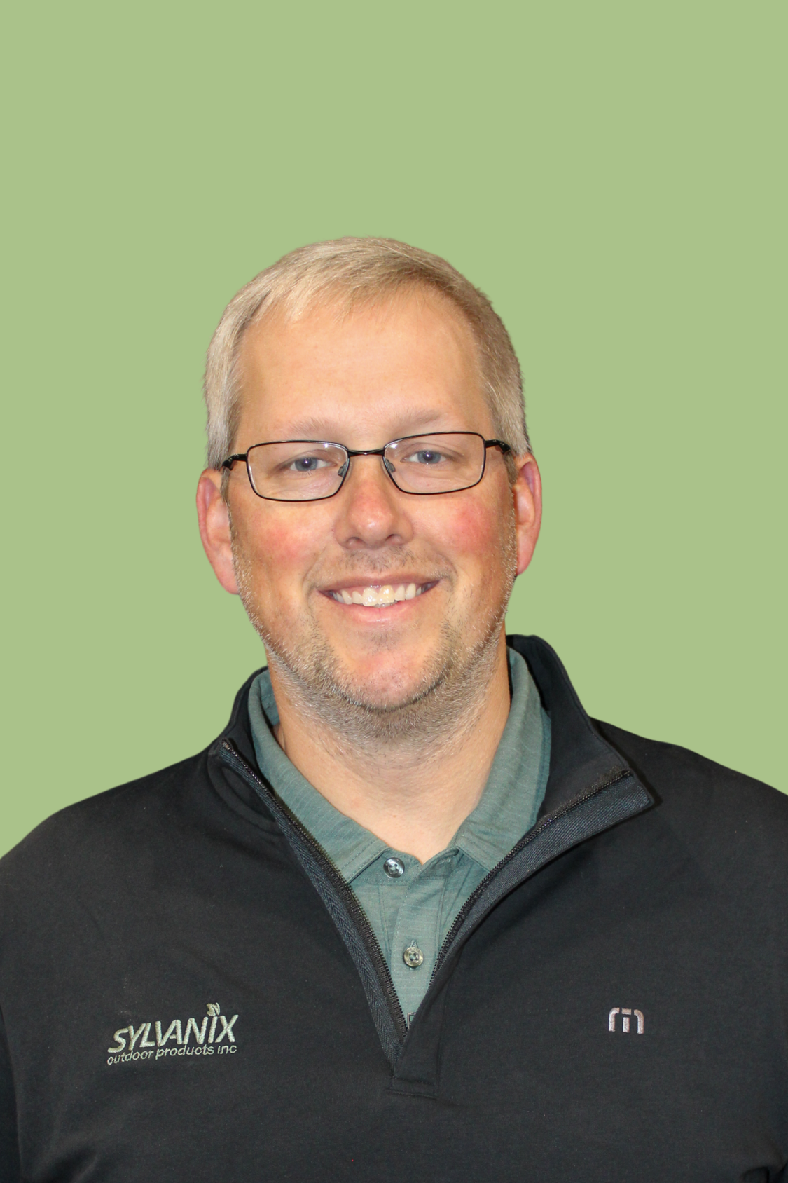 A smiling man with glasses, short blonde hair, and a beard, wearing a gray collared shirt and a black jacket with company logos, standing against a light green background.