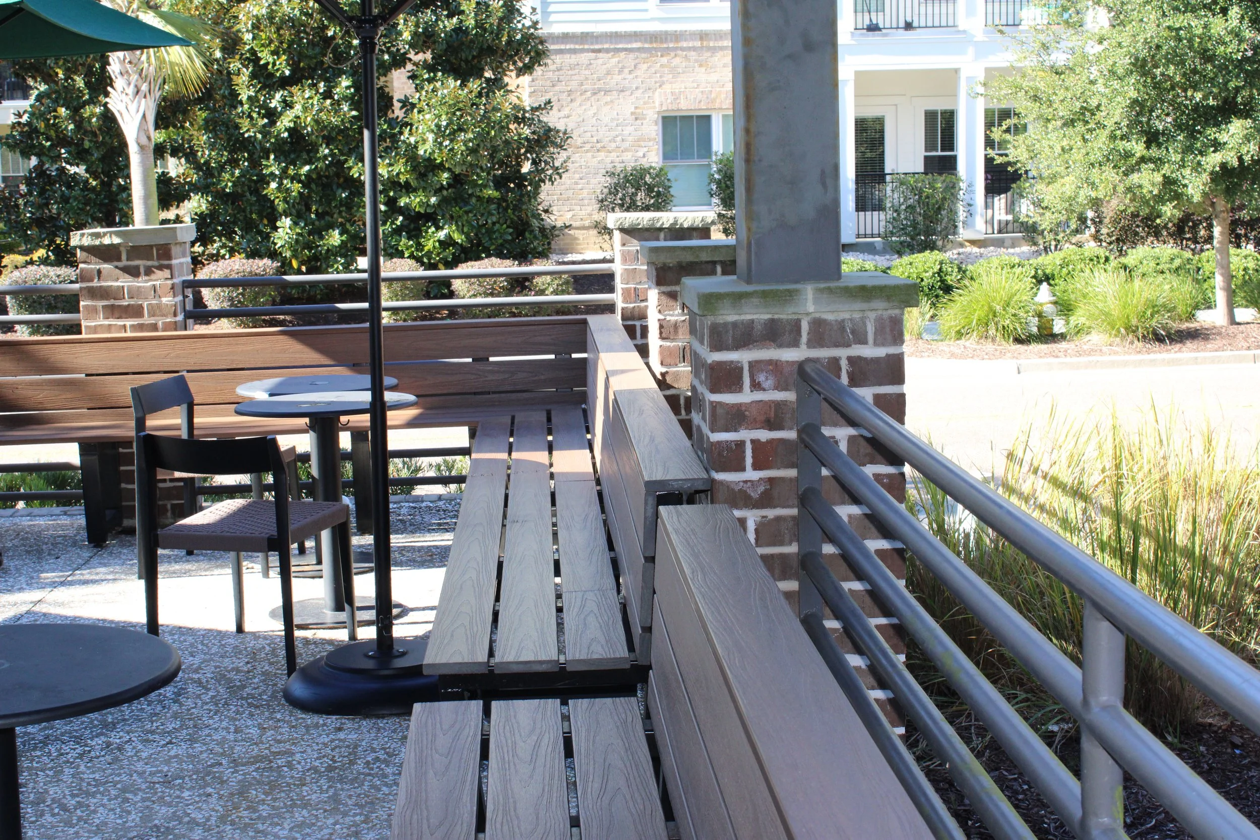 Outdoor patio area with wooden benches, black chairs, tables, and a black umbrella, surrounded by green bushes, trees, and brick railings, with residential buildings in the background.