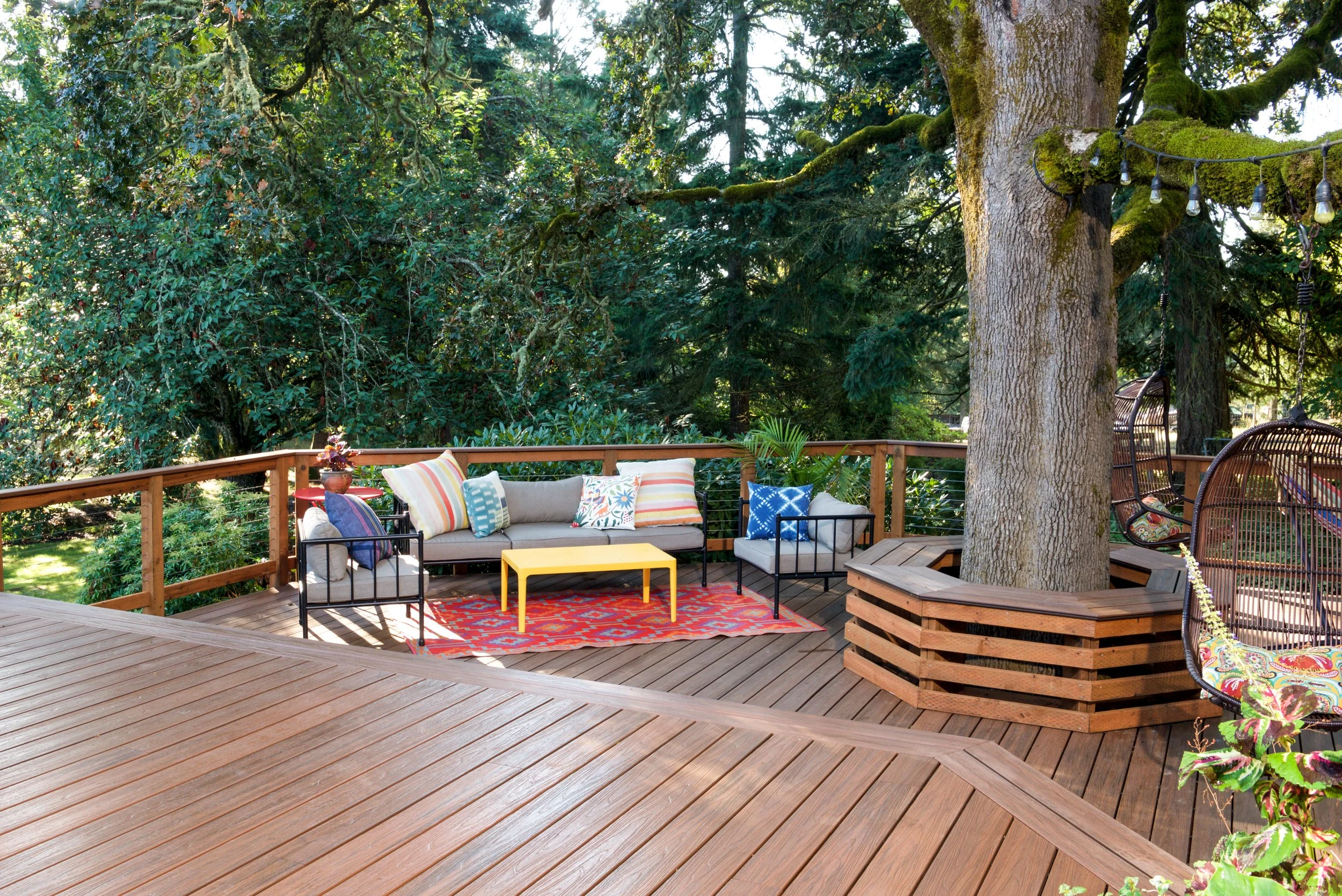 A wooden outdoor deck surrounded by lush green trees. The deck features a couch with colorful pillows, a small yellow table, various chairs, and a string of lights hanging from a large moss-covered tree.