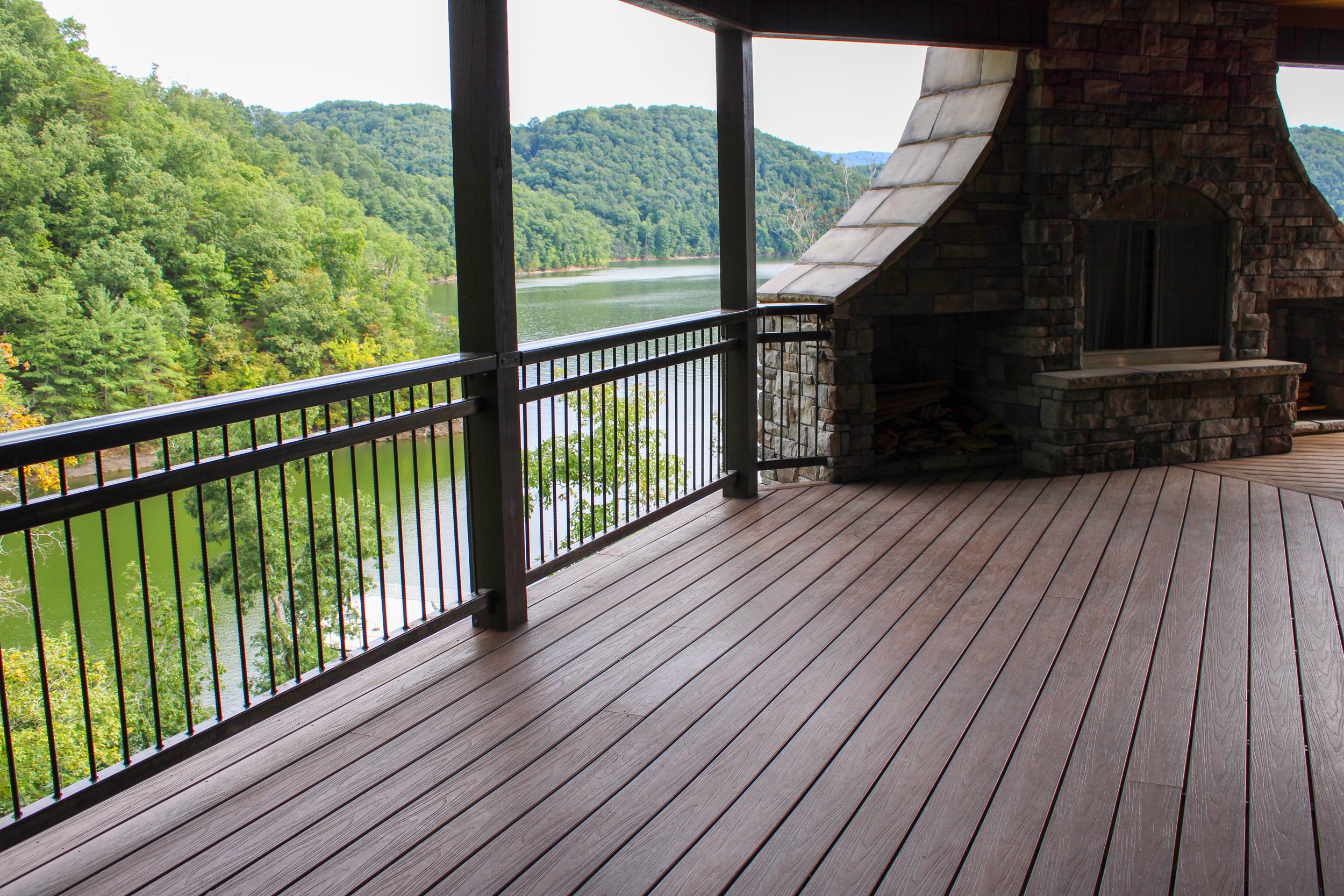 Balcony with wooden floor and iron railing overlooking a river and wooded hills in the distance, with part of a brick and stone outdoor fireplace or grill on the side.
