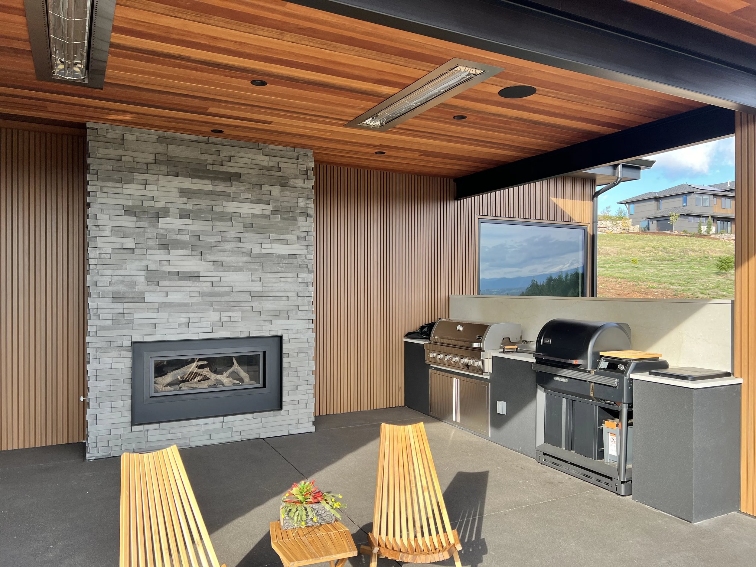Outdoor patio area with a wood ceiling, stone fireplace, and built-in barbecue grill. Two wooden chairs and a small table with a plant are in the foreground, with a view of a house on a hill in the background.