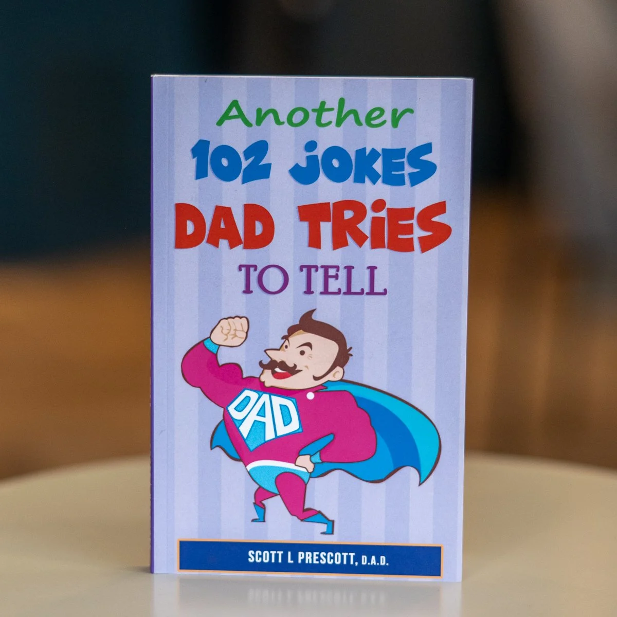 Book titled 'Another 102 Jokes Dad Tries to Tell' with a cartoon illustration of a dad in a superhero costume labeled 'DAD' on the cover.