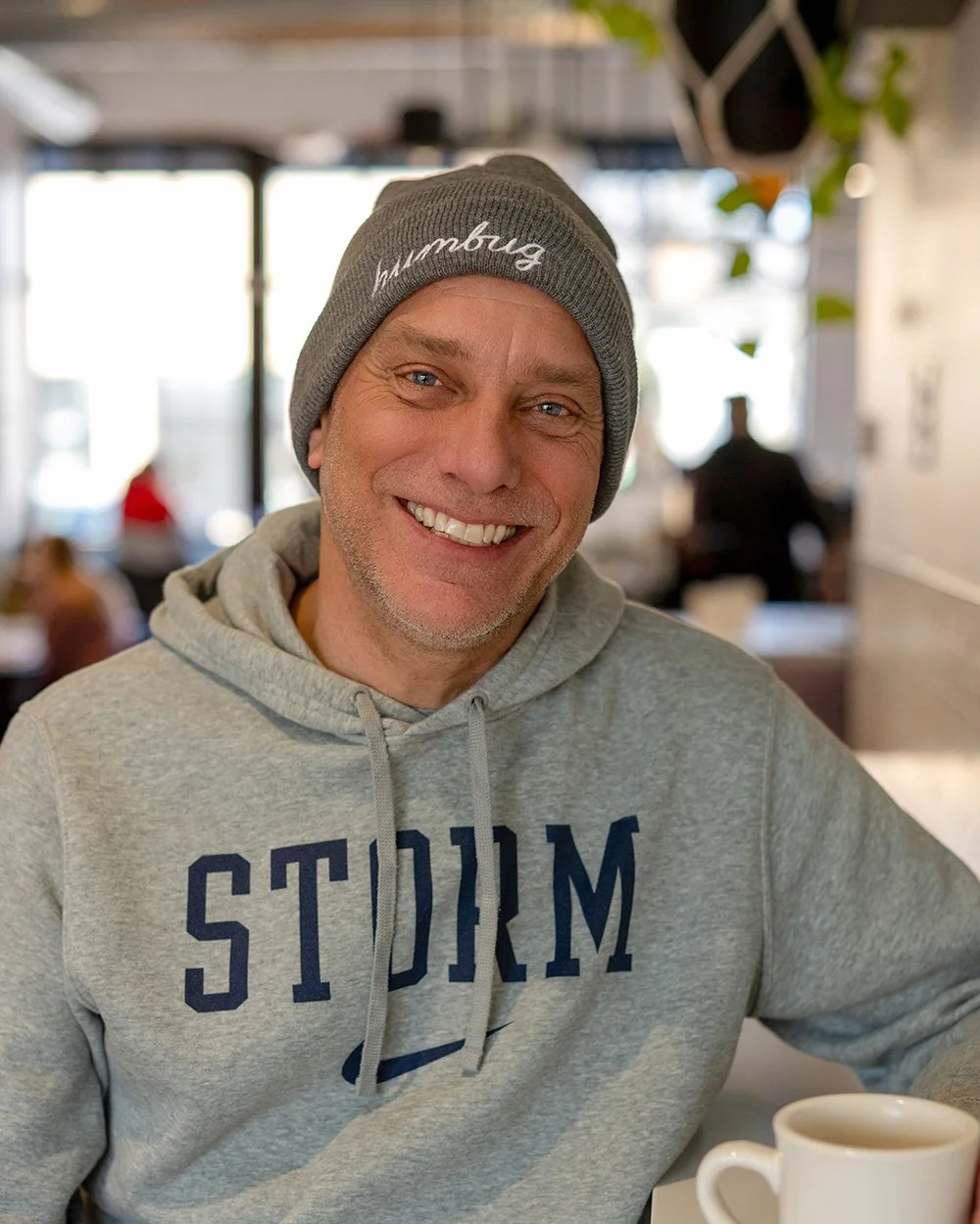 A smiling man wearing a gray hooded sweatshirt and a gray beanie with 'humbug' written on it, sitting at a table in a bright, casual cafe, holding a white mug.