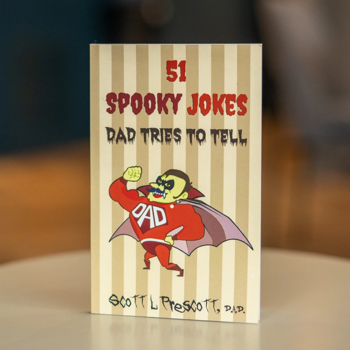 Book titled "51 Spooky Jokes Dad Tries to Tell" by Scott L. Prescott, featuring a cartoon vampire with a "DAD" shirt and a cape, on a beige-striped background.