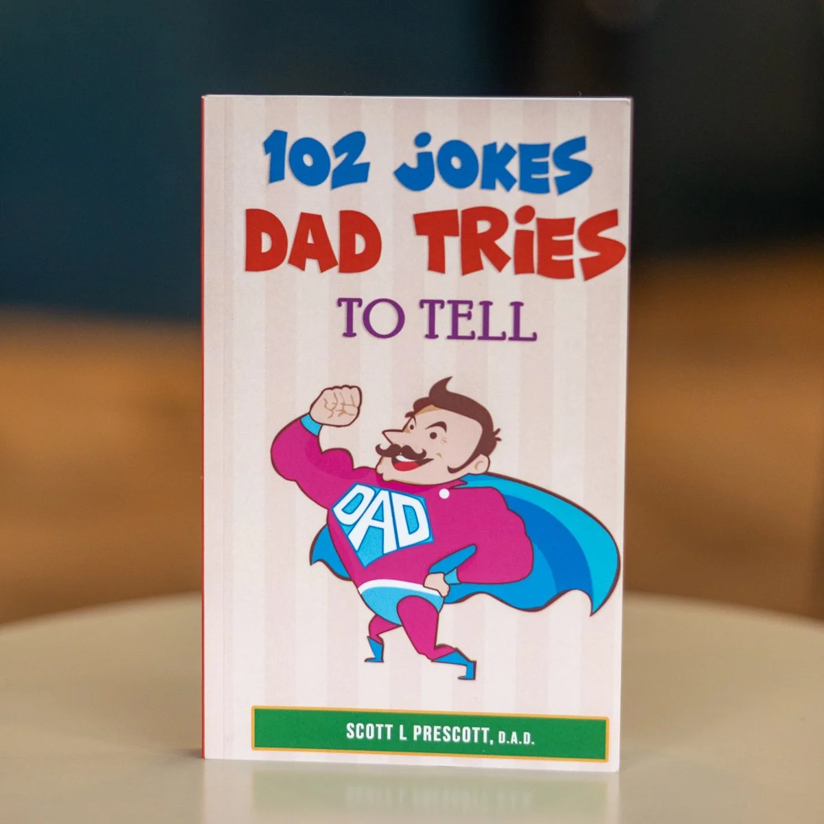 Book titled "102 Jokes Dad Tries To Tell" by Scott L Prescott, featuring a cartoon of a man dressed as a superhero with "DAD" on his chest, flexing his arm.