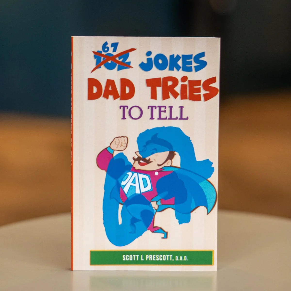Book titled "67 Jokes Dad Tries to Tell" by Scott L. Prescott, D.A.D., with a cartoon illustration of a superhero dad in a blue cape and costume fighting a blue creature.