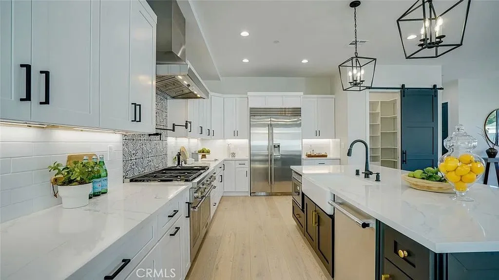 Modern kitchen with white cabinets, stainless steel appliances, and a large island. Features include a stovetop with range hood, pendant lighting, a farmhouse sink, decorative backsplash, and bowls of fruit on the island.