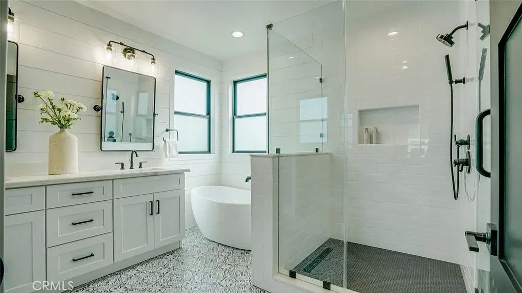 Modern bathroom with a freestanding bathtub, glass-enclosed shower, double vanity with black fixtures, and decorative floor tiles.