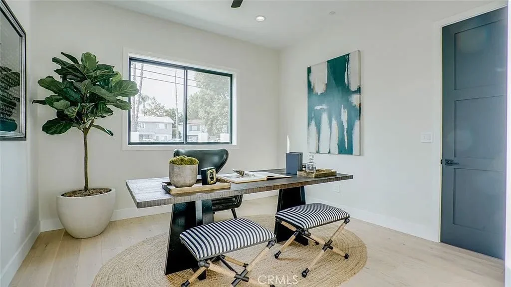 Modern home office with a wooden desk, black leather chair, potted plant, abstract artwork, striped stools, and glass window.