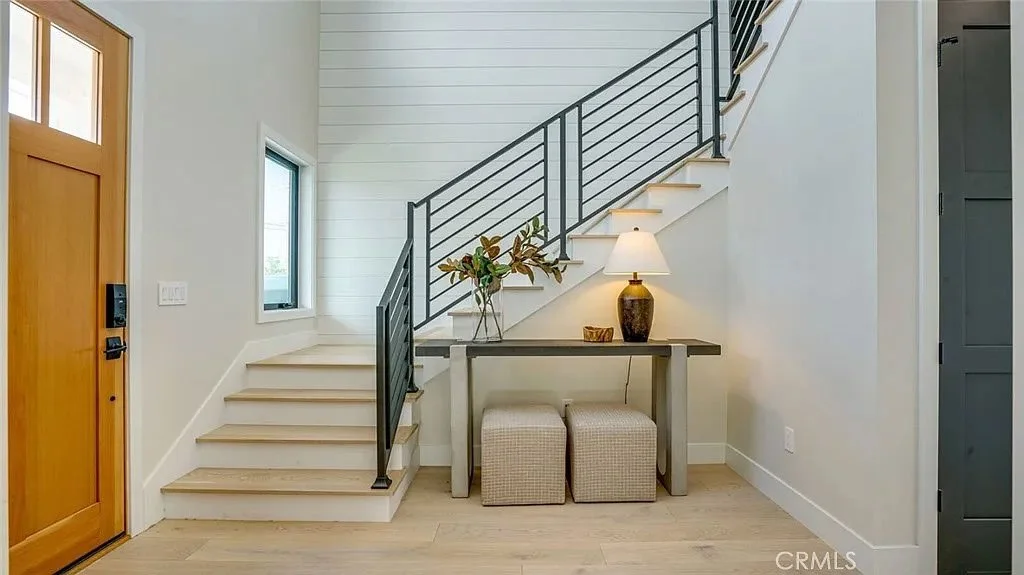 Modern entryway with staircase, wooden door, lamp, table, and two ottomans.