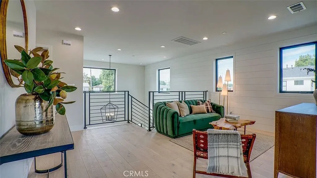 Modern living room with green sofa, decorative pillows, and floor lamp. Features a wooden chair with plaid throw, wooden coffee table, and large windows. Includes a console table with a potted plant and round mirror.