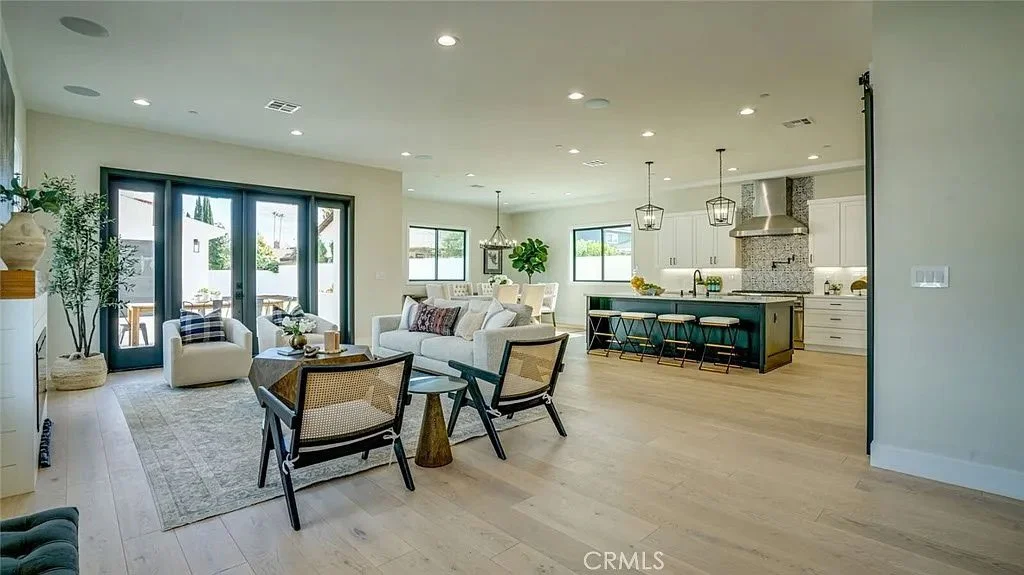 Modern open-concept living room and kitchen with light wood flooring, white walls, recessed lighting, a large black-framed glass door, white sofas, a dark kitchen island with bar stools, and contemporary pendant lights.