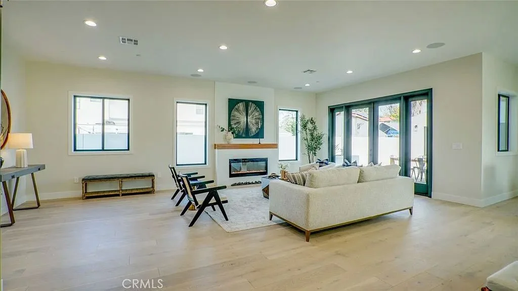 Modern living room with hardwood floors, light-colored walls, and recessed lighting. Features a sofa, armchairs, fireplace, and large windows with view of the outdoor area.