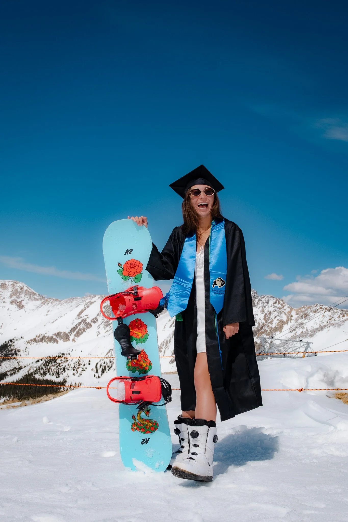 A woman in a graduation gown and cap, holding a snowboard with red bindings, standing on snow-covered ground in a mountainous area with snow-capped peaks and a clear blue sky.