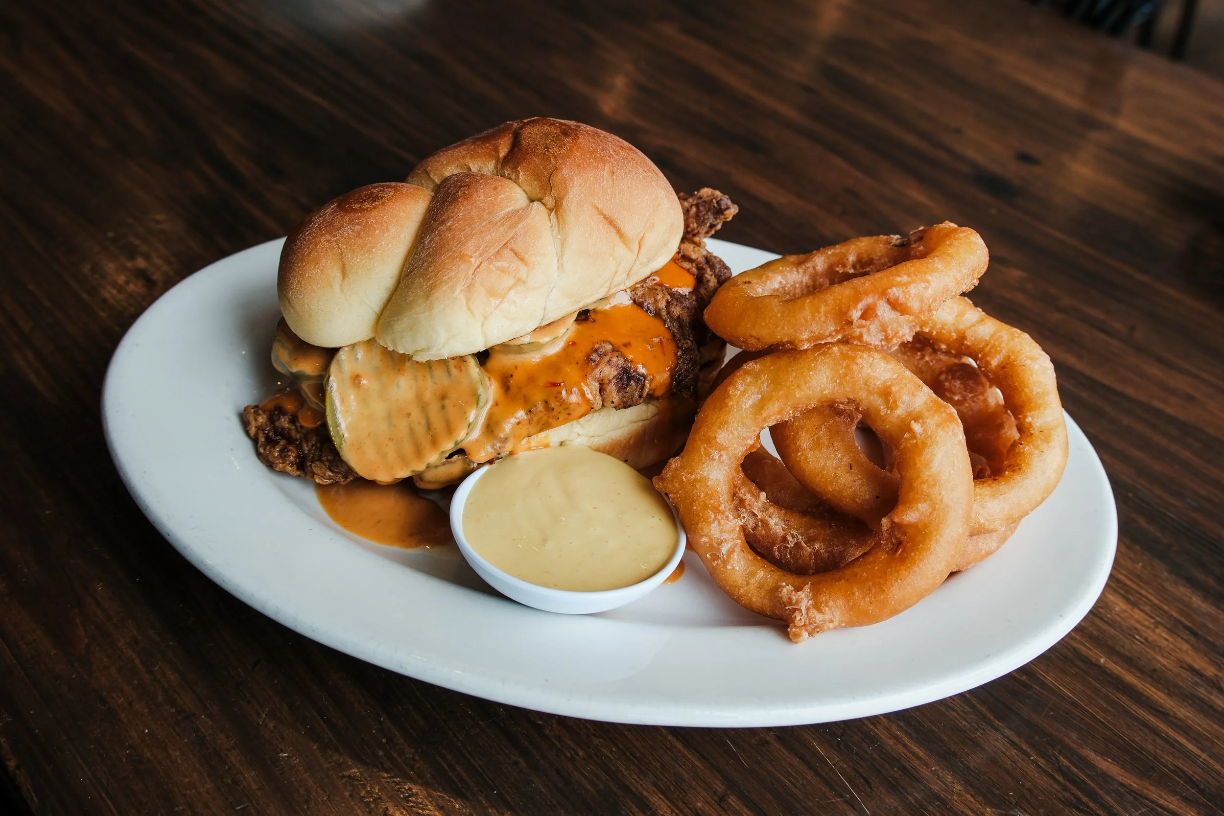 Fried chicken sandwich with onion rings and dipping sauces on a white plate.