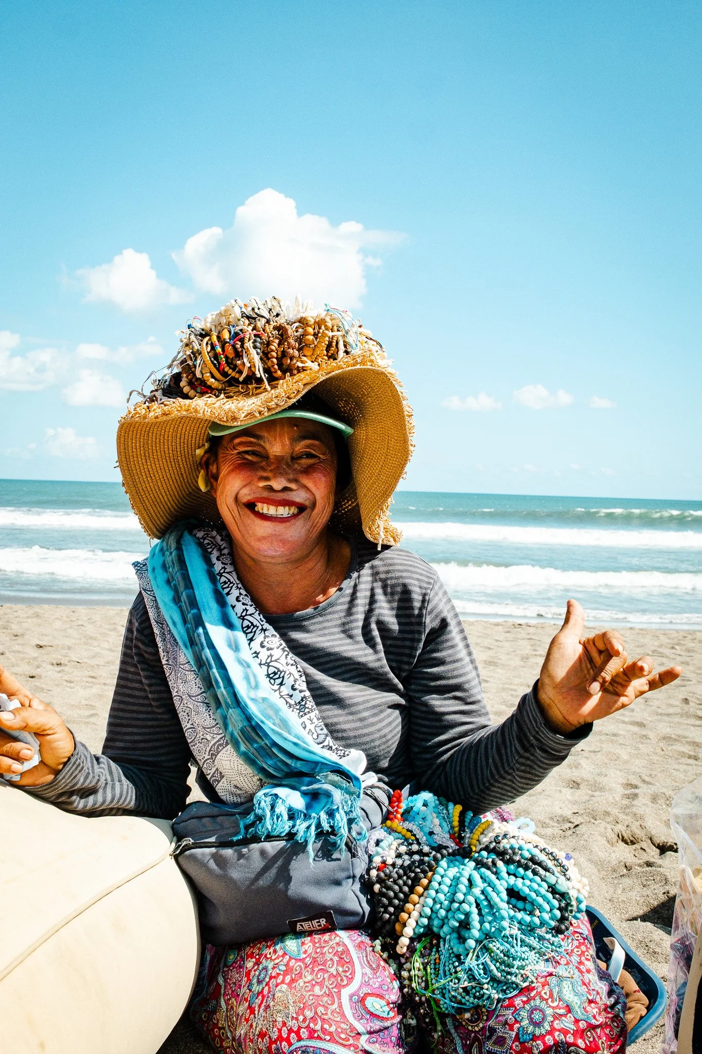 A smiling woman sitting on the beach wearing a large straw hat adorned with jewelry and a scarf, with a pile of necklaces beside her, and the ocean and blue sky with clouds in the background.