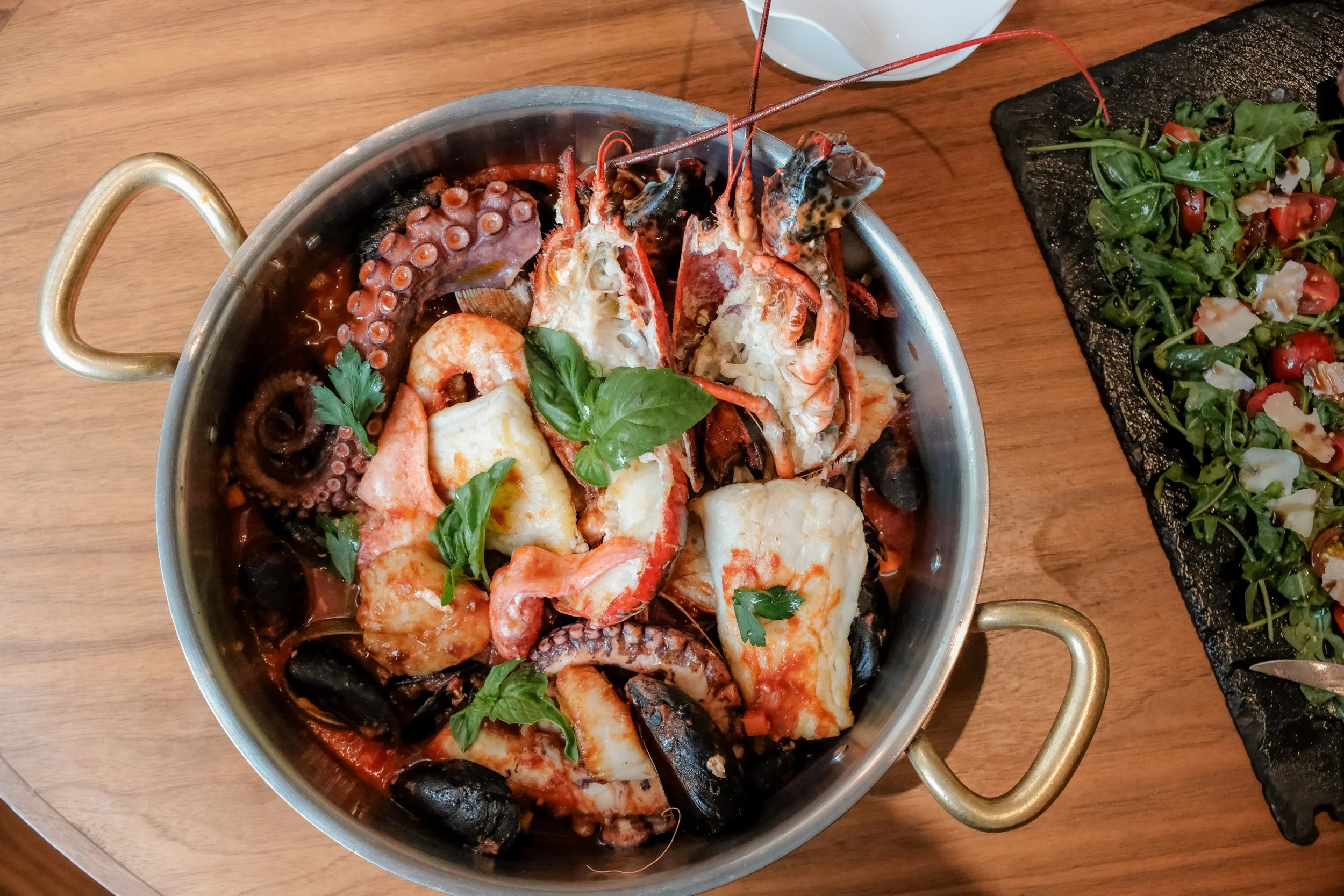 Seafood stew with lobster, shrimp, octopus tentacles, mussels, and fresh basil in a metal pot on a wooden table.