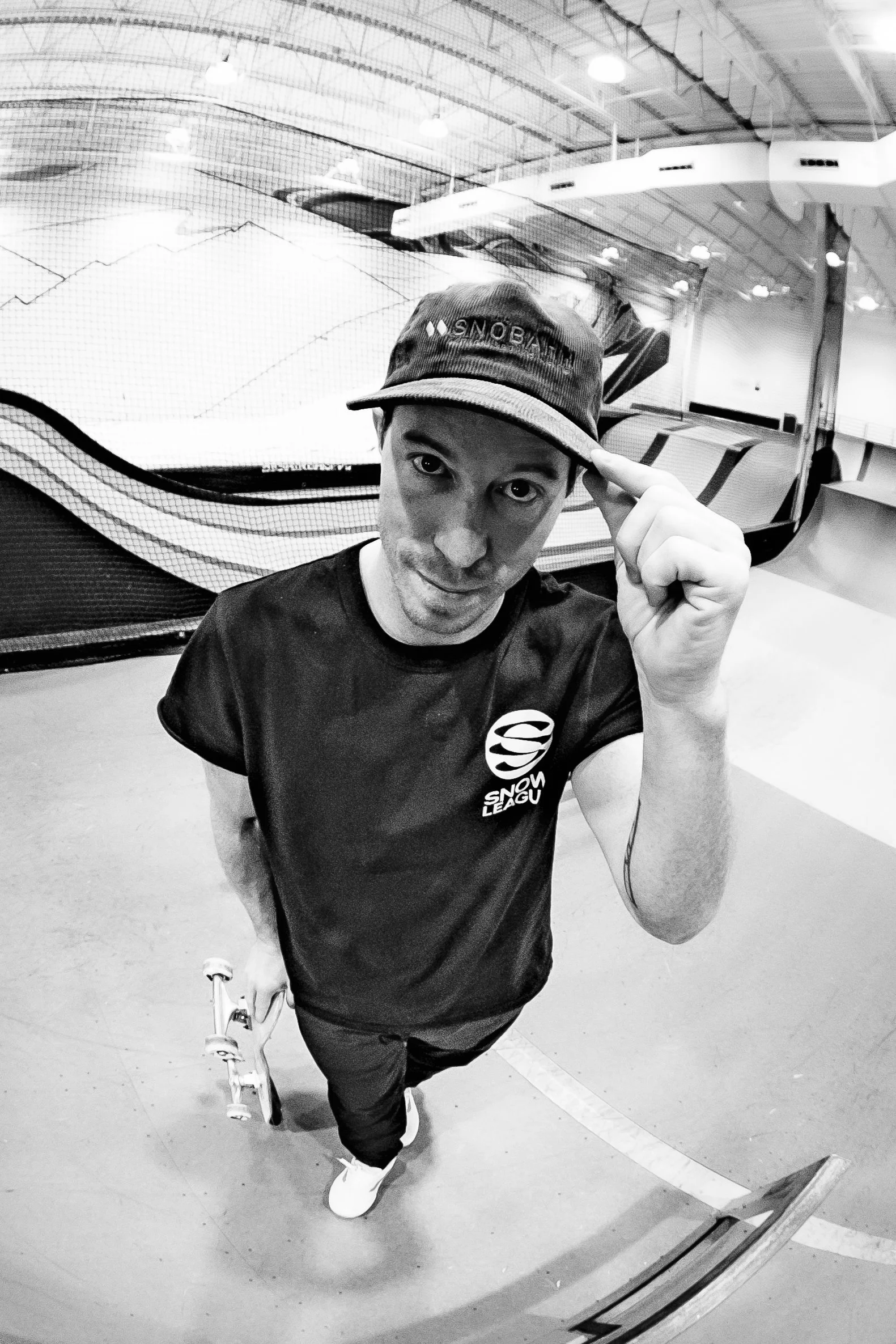 Shawn White, wearing a black T-shirt with a snow league logo, a cap that says 'SNOBAHN,' and holding a skateboard, is taking a selfie inside an indoor skate park.