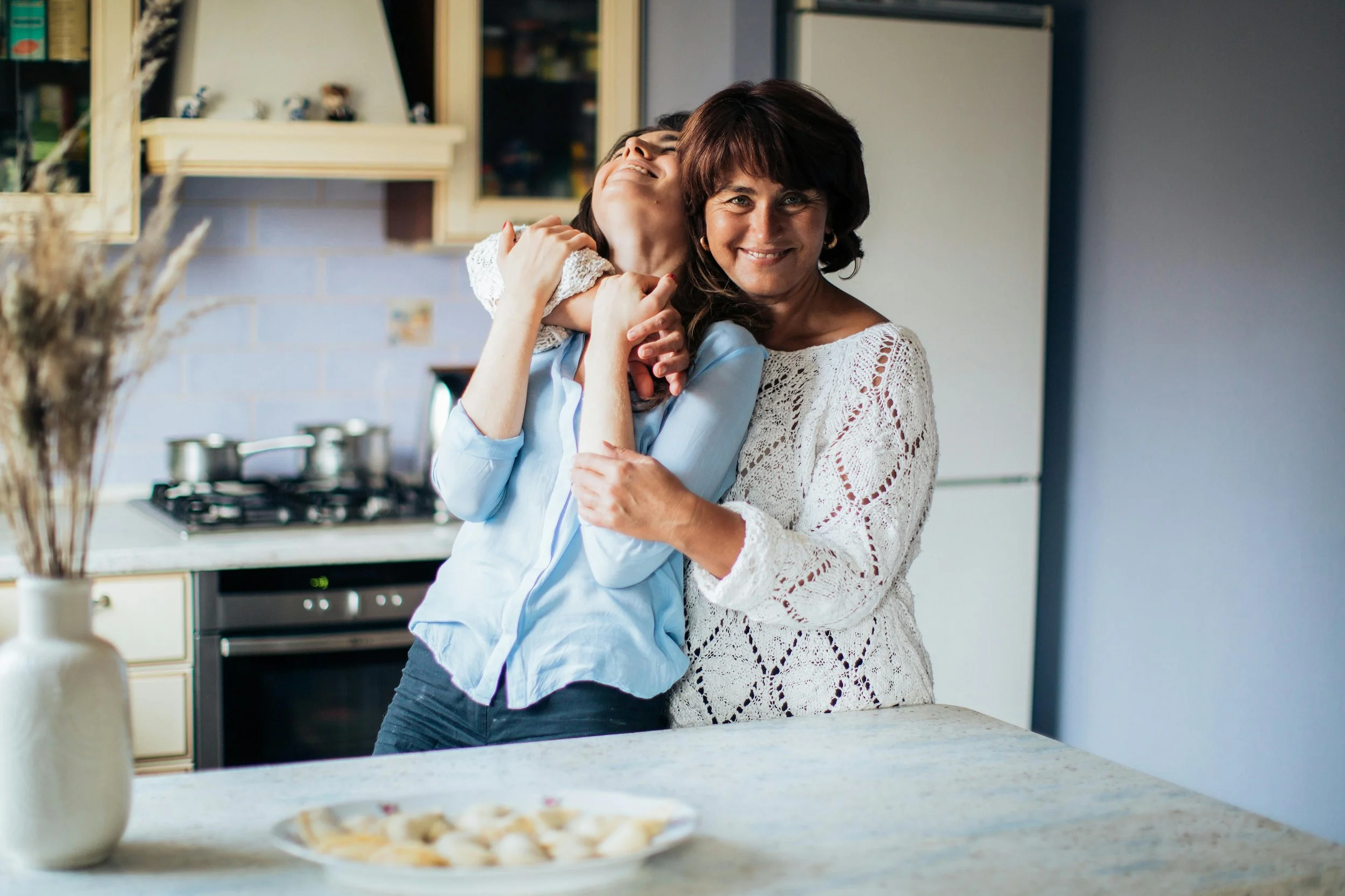 Mother and adult daughter in a playful hug in the kitchen, with a tray of cookies nearby