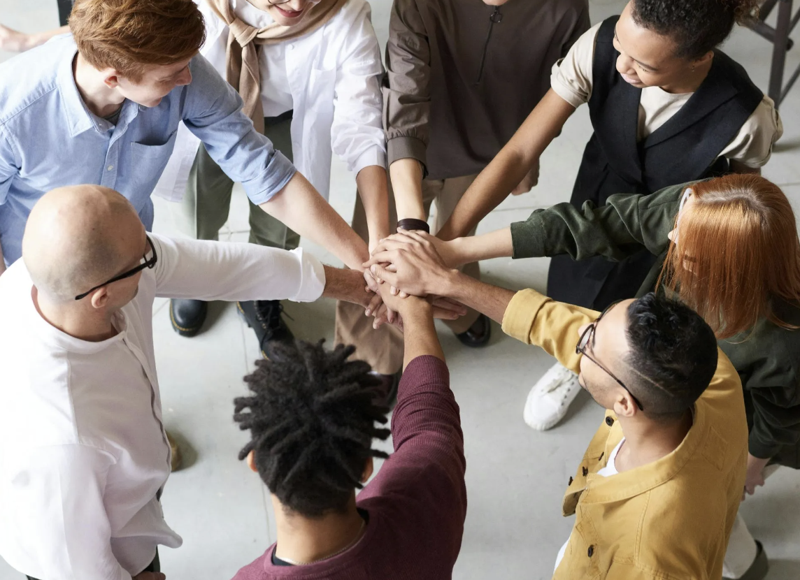 A diverse group of eight people standing in a circle, putting their hands together in the middle for a group cheer or show of unity.
