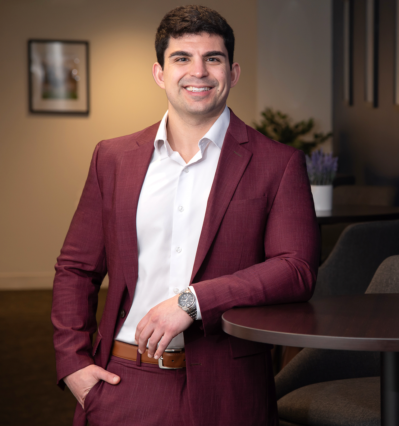 A young man in a burgundy suit with a white shirt, smiling, leaning on a round table in an indoor setting.