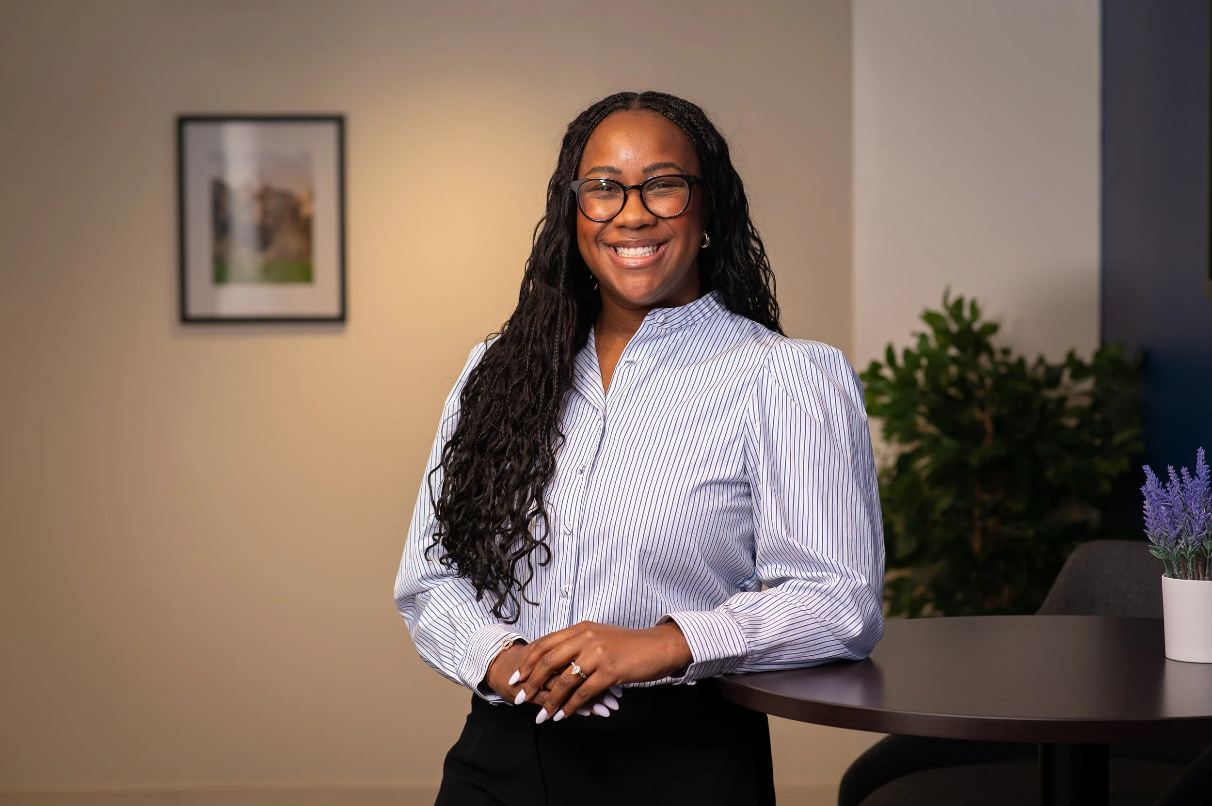 A woman with glasses and natural curly hair smiling at the camera in front of a plain light-colored wall.
