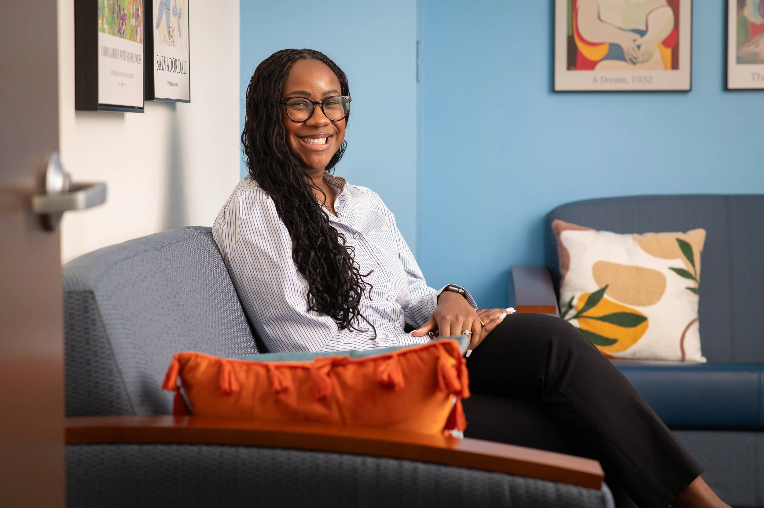 A woman with glasses and natural curly hair smiling at the camera in front of a plain light-colored wall.