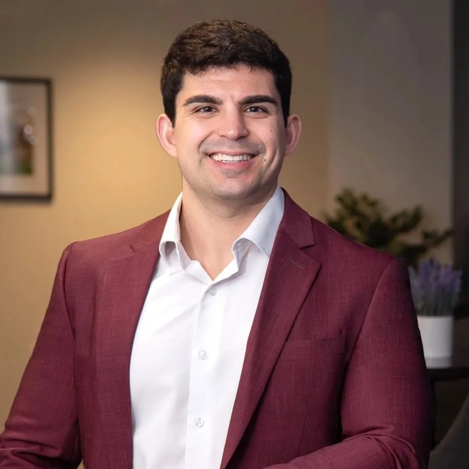 A young man in a burgundy suit with a white shirt, smiling, leaning on a round table in an indoor setting.