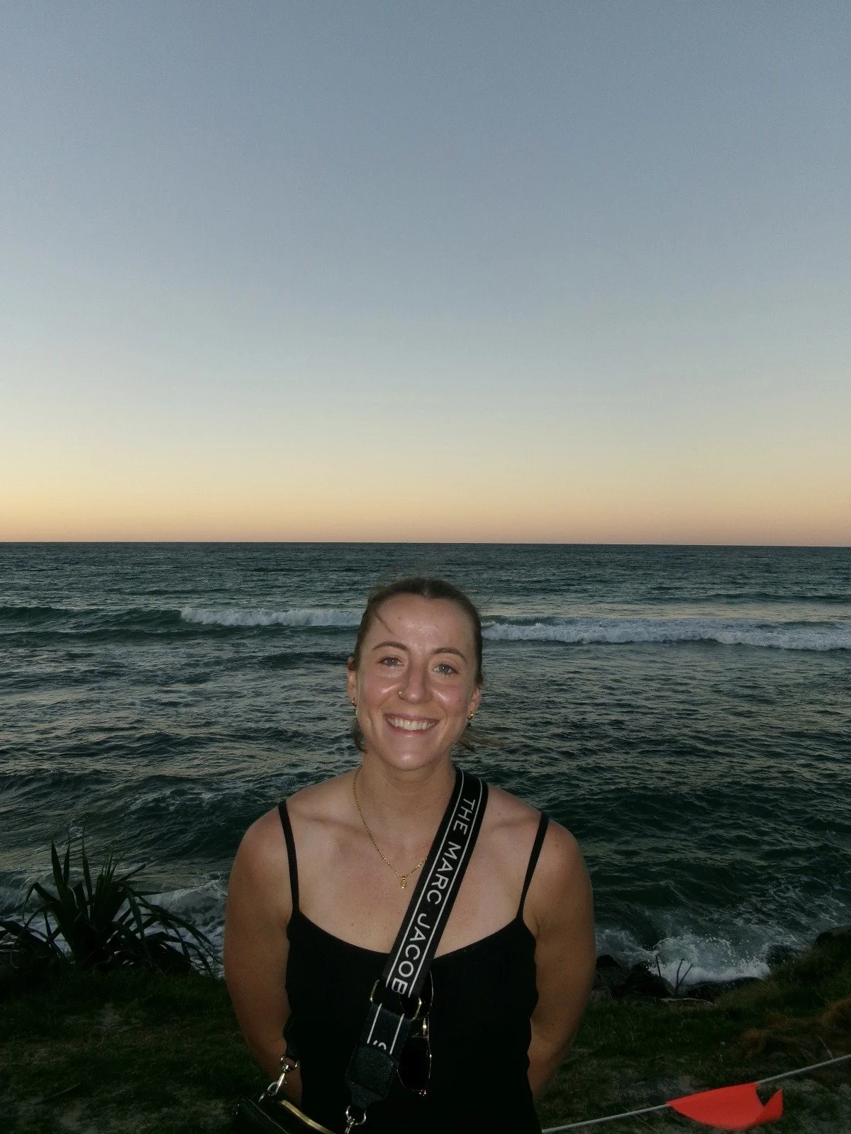 A woman smiling at the camera standing in front of the ocean during sunset, wearing a black top and a camera strap around her neck.