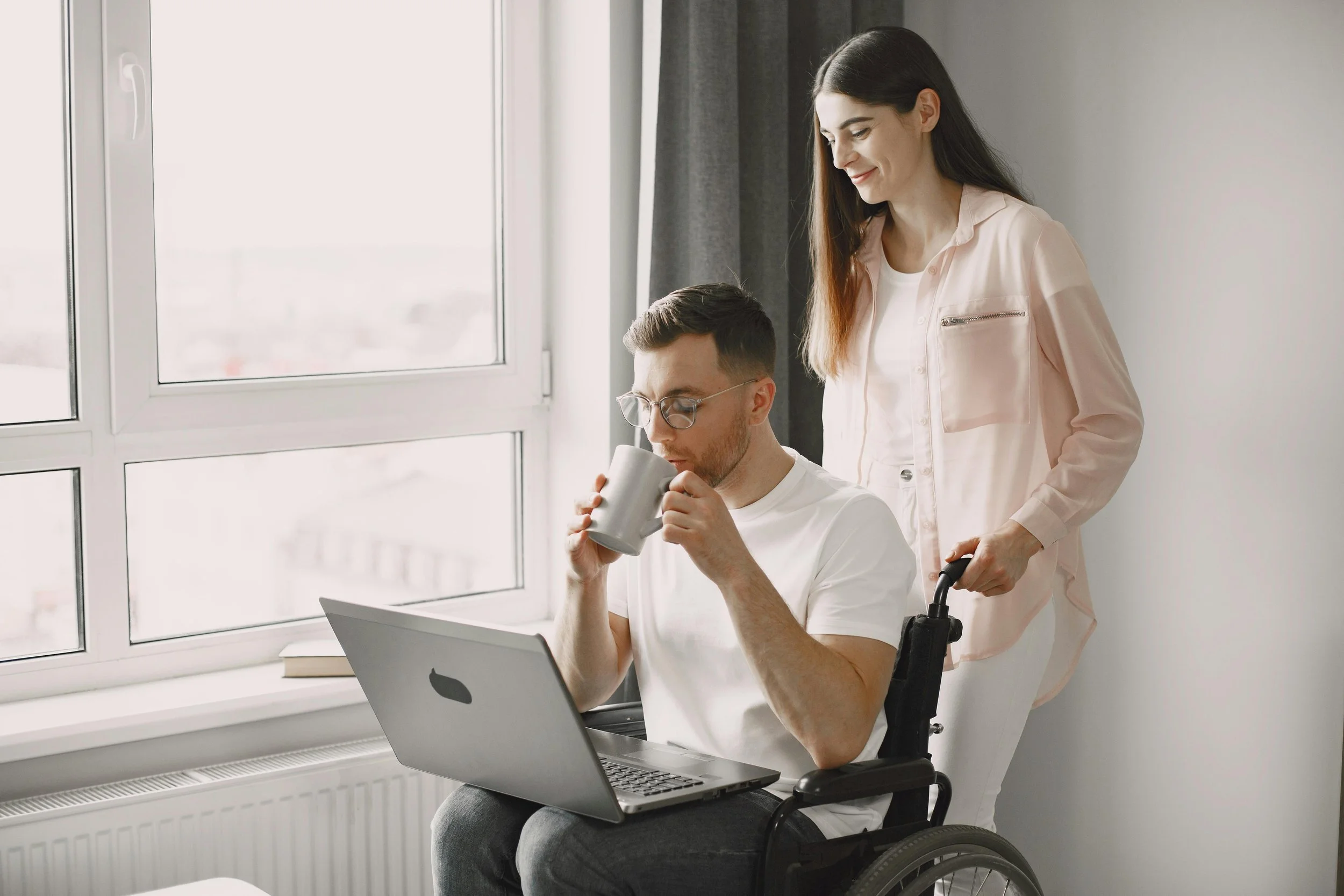 A man in a wheelchair drinking from a mug while looking at a laptop, with a woman standing behind him pushing his wheelchair, in a brightly lit room near a large window.