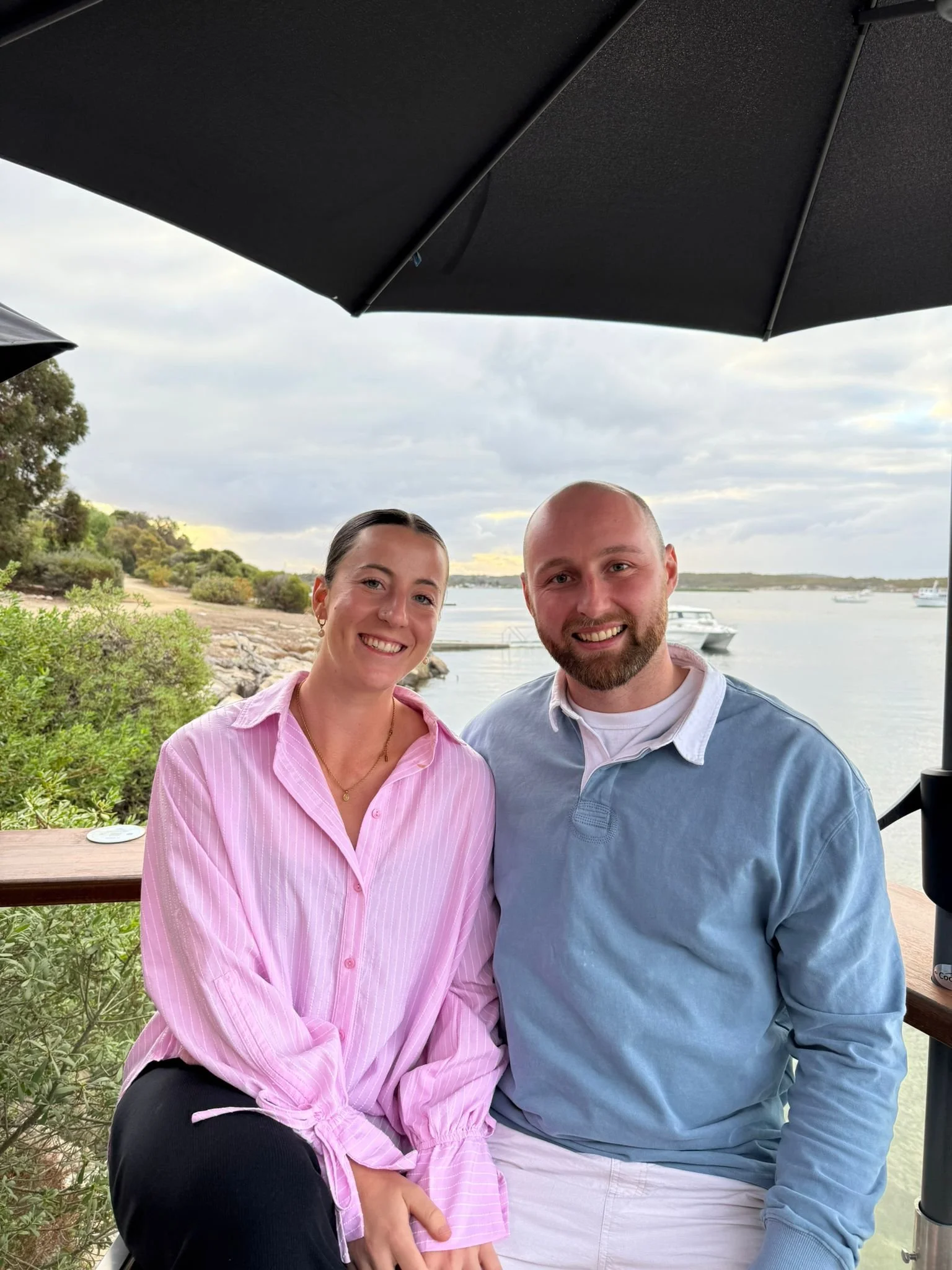 A smiling couple sitting outdoors near a body of water under a large black umbrella, with boats and greenery in the background.