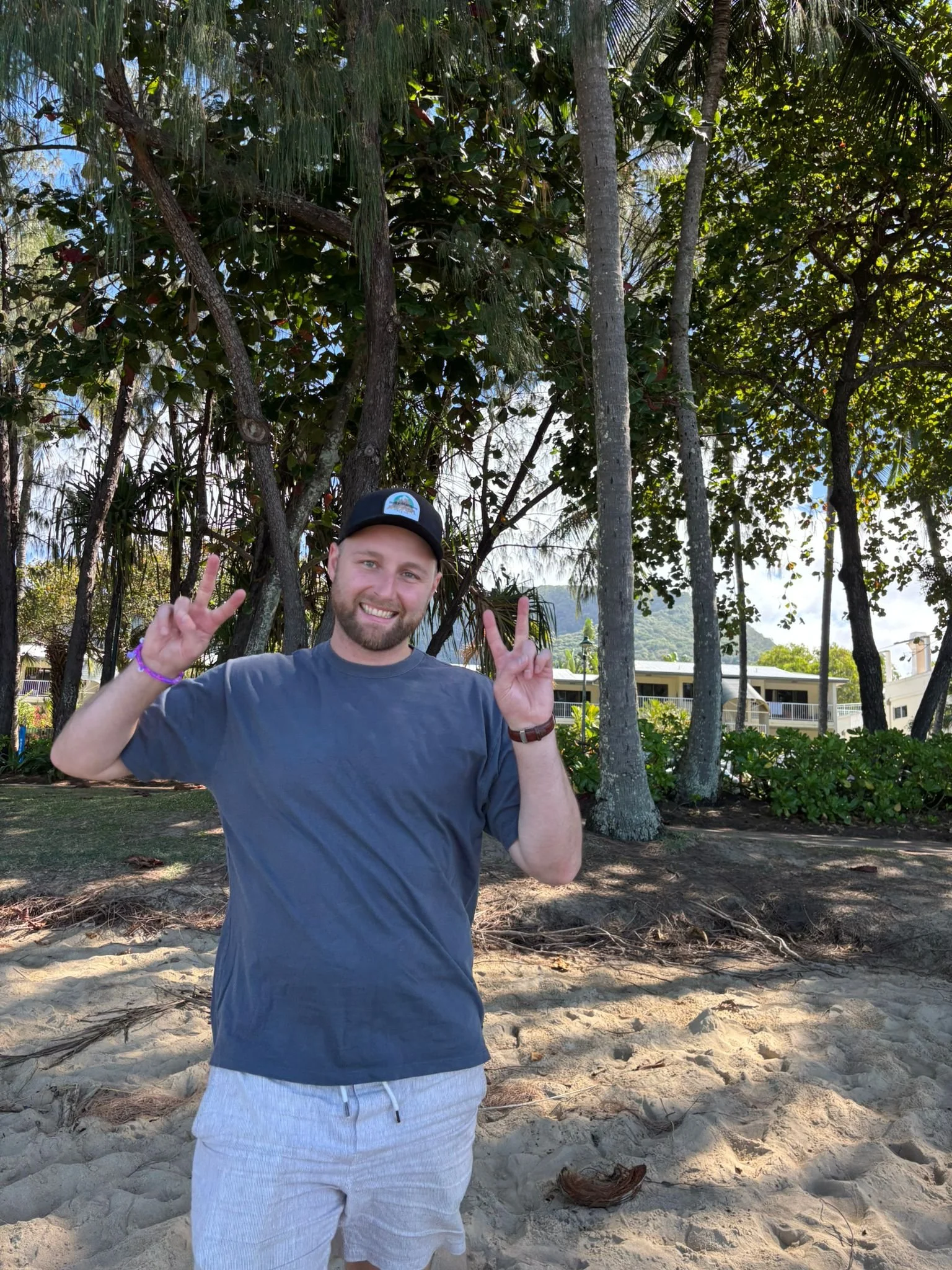 A man smiling and making peace signs with both hands standing on a sandy beach with trees and buildings in the background.