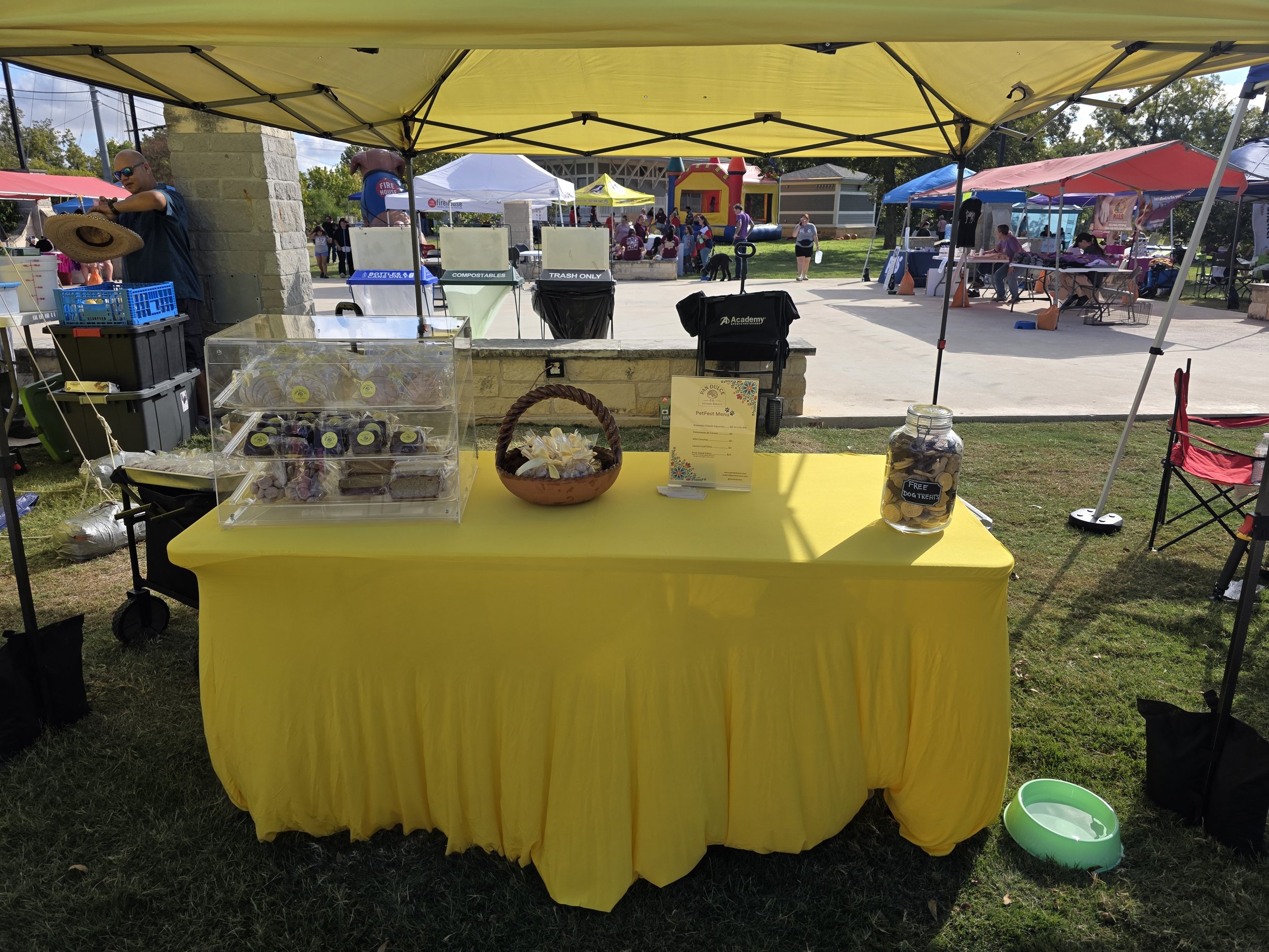 Pan Dulce Co. first booth with a yellow tablecloth at an outdoor event, offering dog treats in a jar, with a sign and a basket, under a yellow canopy, with people, tents, and activities visible in the background.