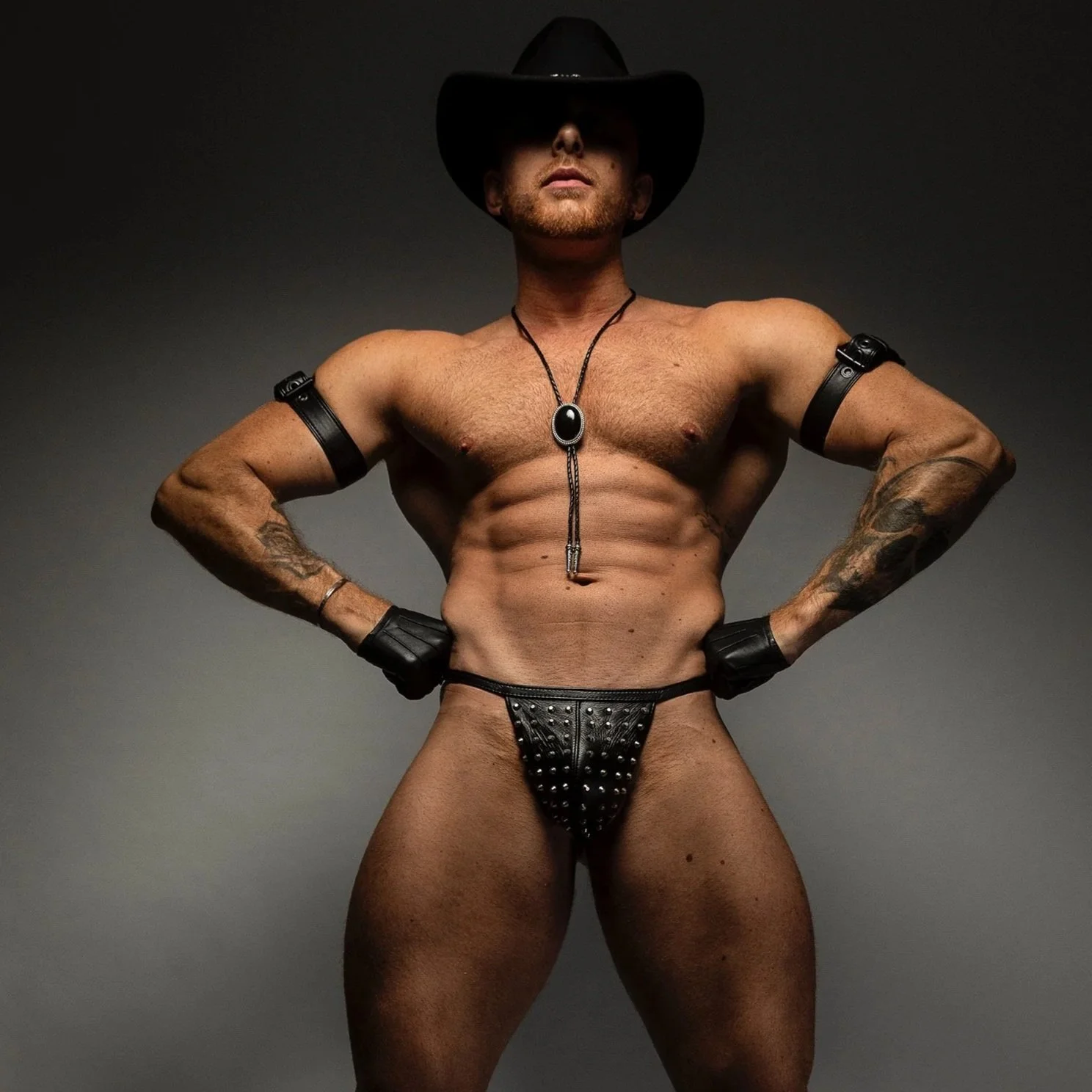A muscular shirtless man wearing a black cowboy hat, black gloves, and studded black underwear, with his hands on his hips, against a plain dark background.