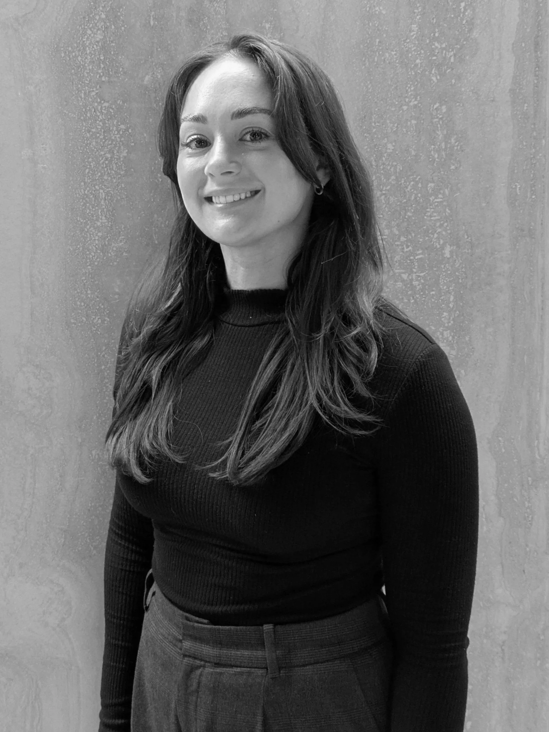 Black and white photo of a woman smiling, wearing a black top and plaid pants, standing against a plain textured background.