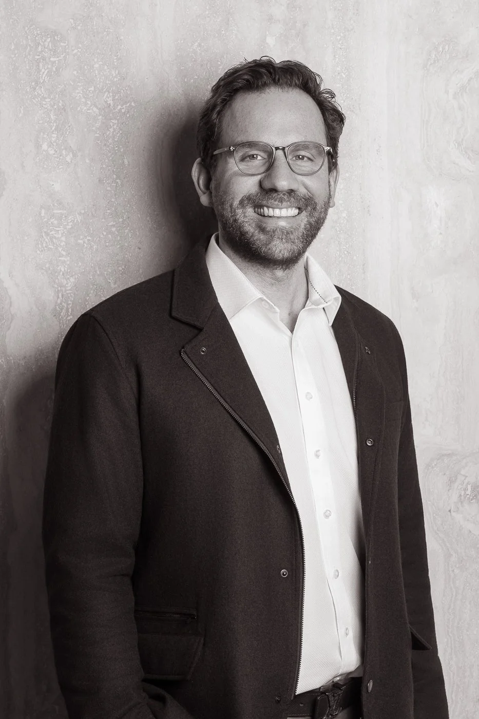 Black and white photo of a man with glasses, a beard, and curly hair, smiling while leaning against a textured wall. He is wearing a white button-up shirt and a dark blazer.