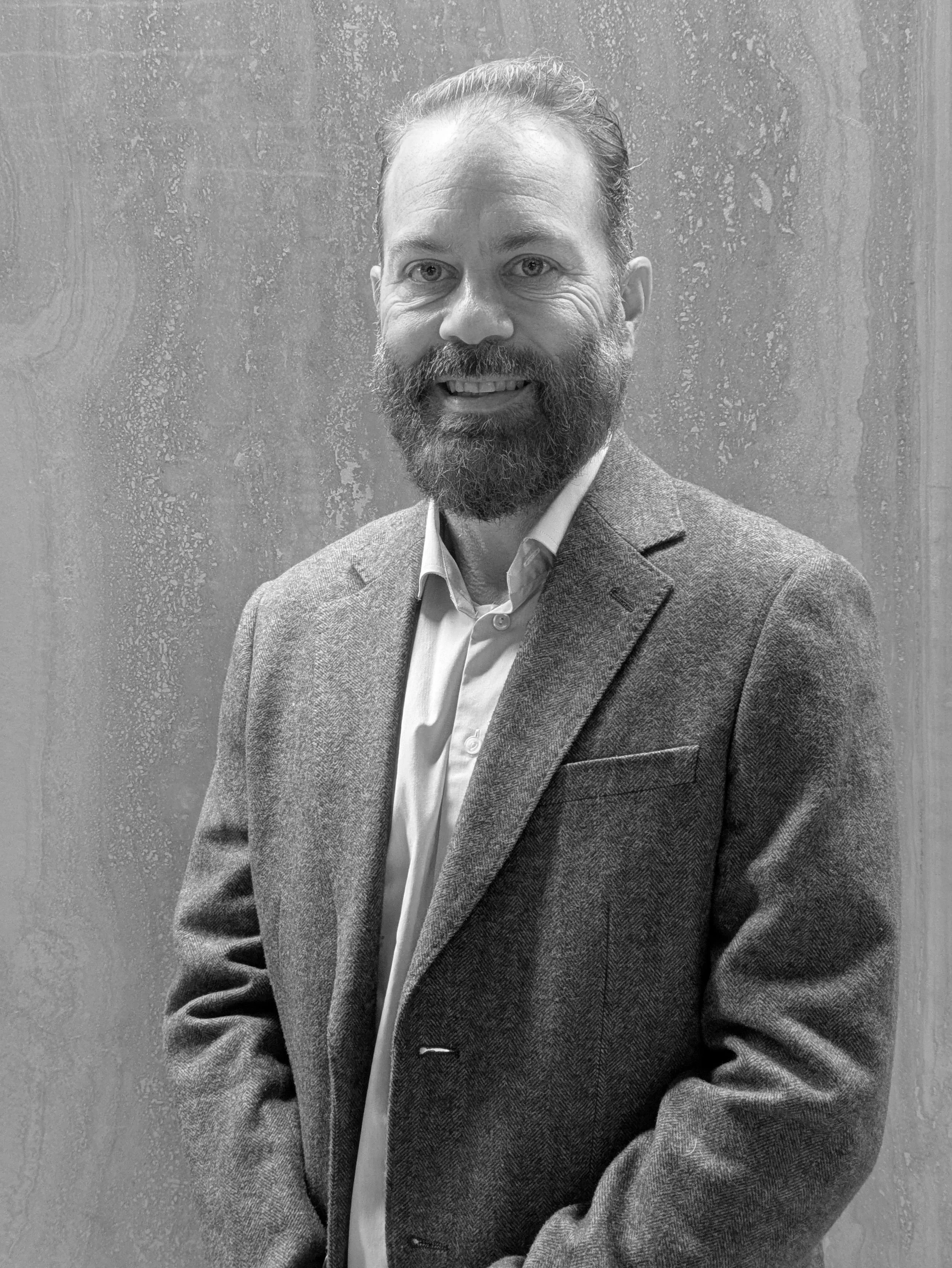 Black and white photo of a man with a beard, wearing a blazer and collared shirt, smiling, against a textured background.