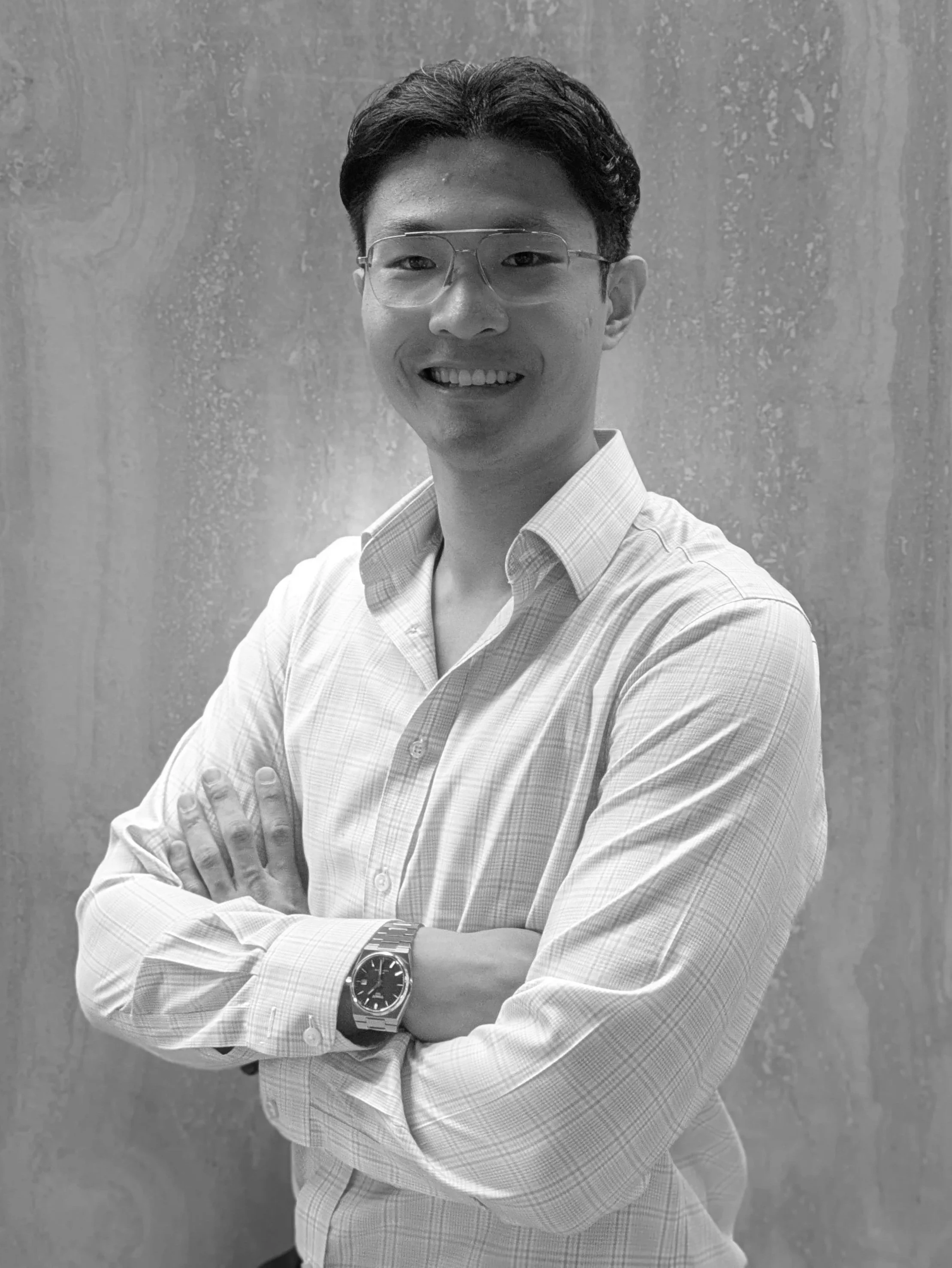 A young man smiling, wearing glasses, a light-colored checkered shirt, a wristwatch, and a bracelet, standing with arms crossed against a textured wall background.